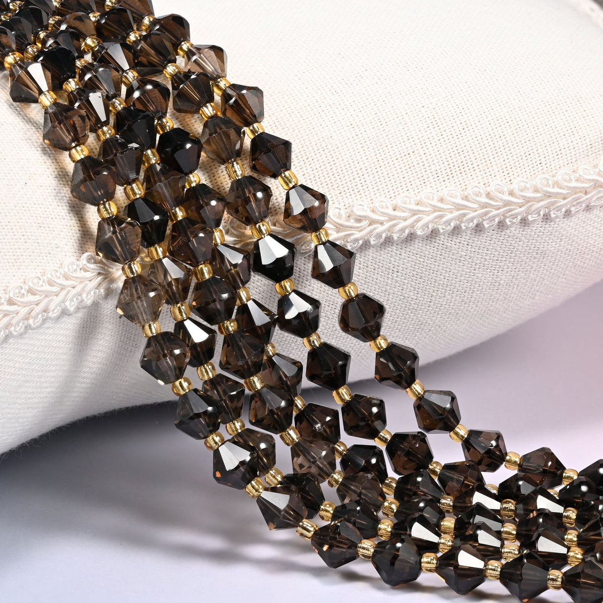 Smoky Quartz faceted bicone beads, 8mm, draped diagonally over a white fabric cushion showing the warm brown-gray translucent tone.