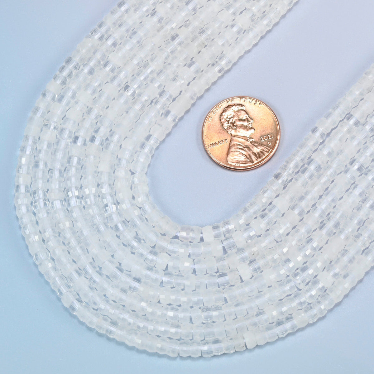 Close view of clear quartz faceted heishi disc strands curving beside a US penny for scale