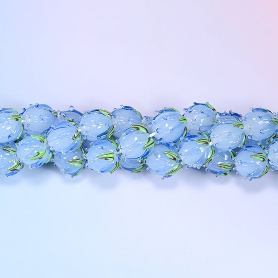 Blue tulip buds worked by hand at the lamp — closed petals sculpted into the glass and a green leaf detail wrapped at the base, so each bead reads as a small flower rather than a flower-shaped disc.