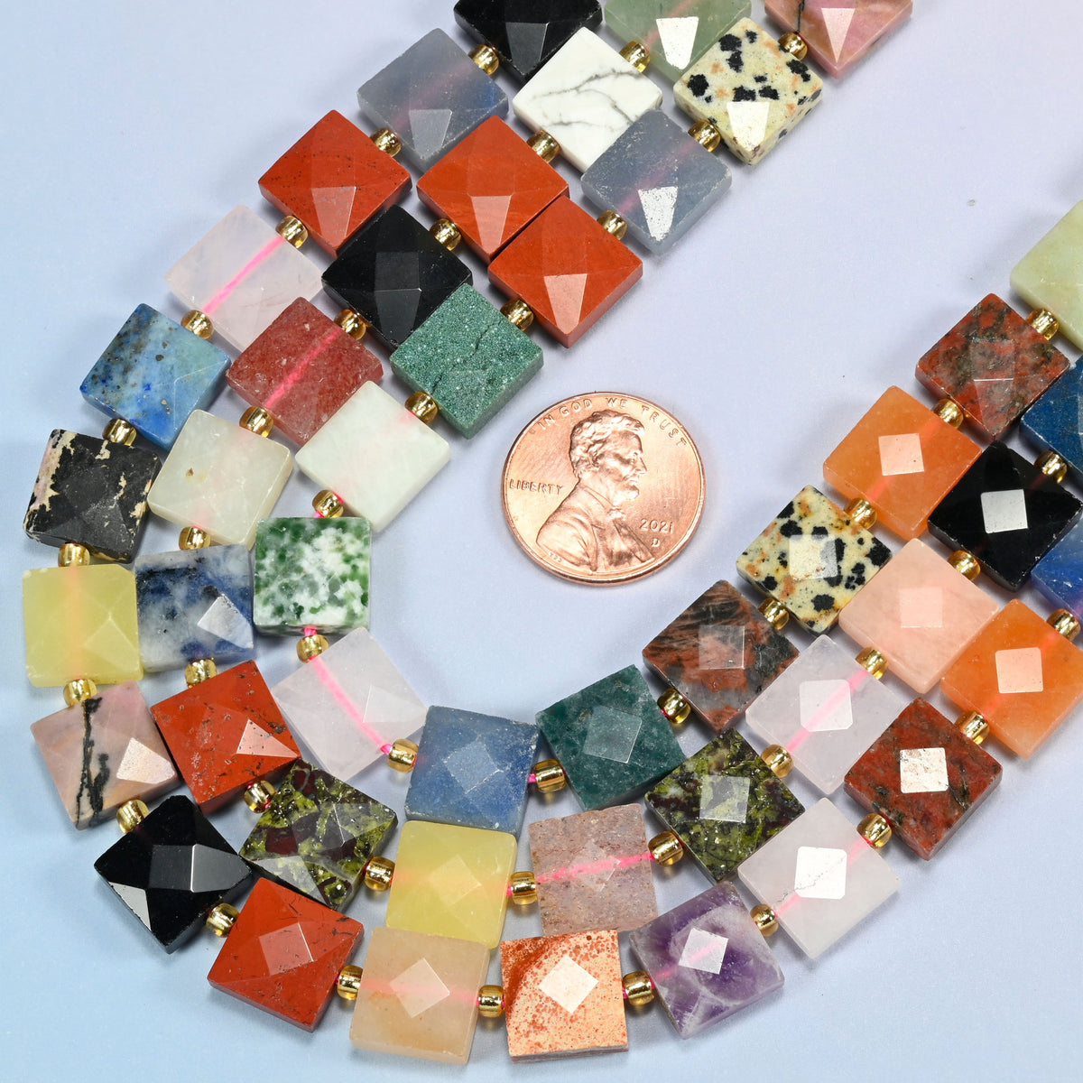 Close view of assorted gemstone faceted flat square strands curving beside a US penny for scale