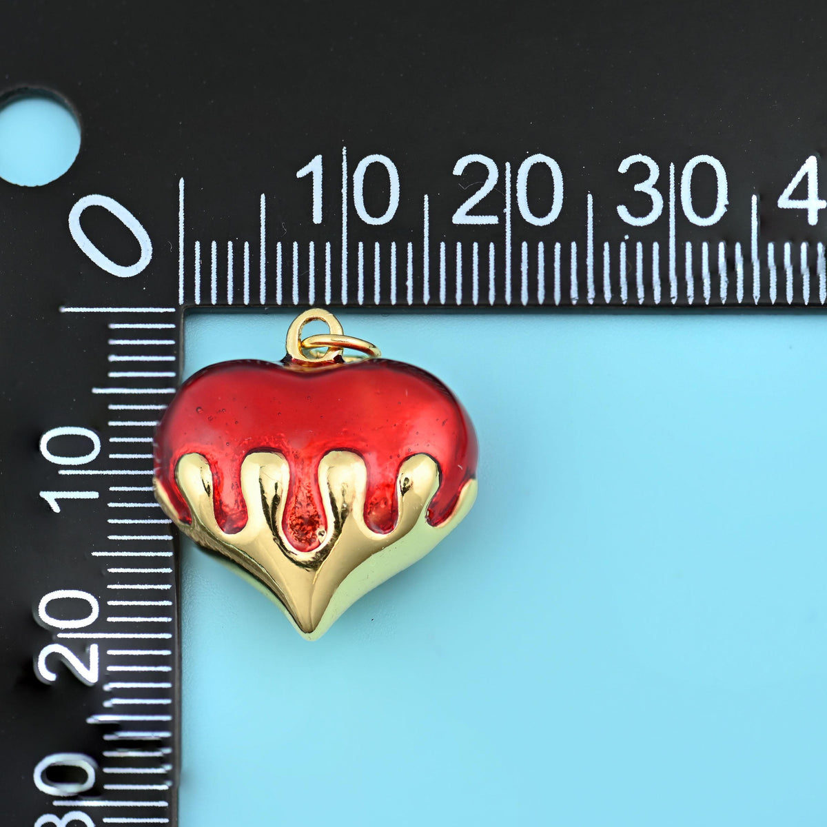 Enamel dripping puffy heart pendant in red measured on a black millimetre ruler, the charm reaching about 20mm across.