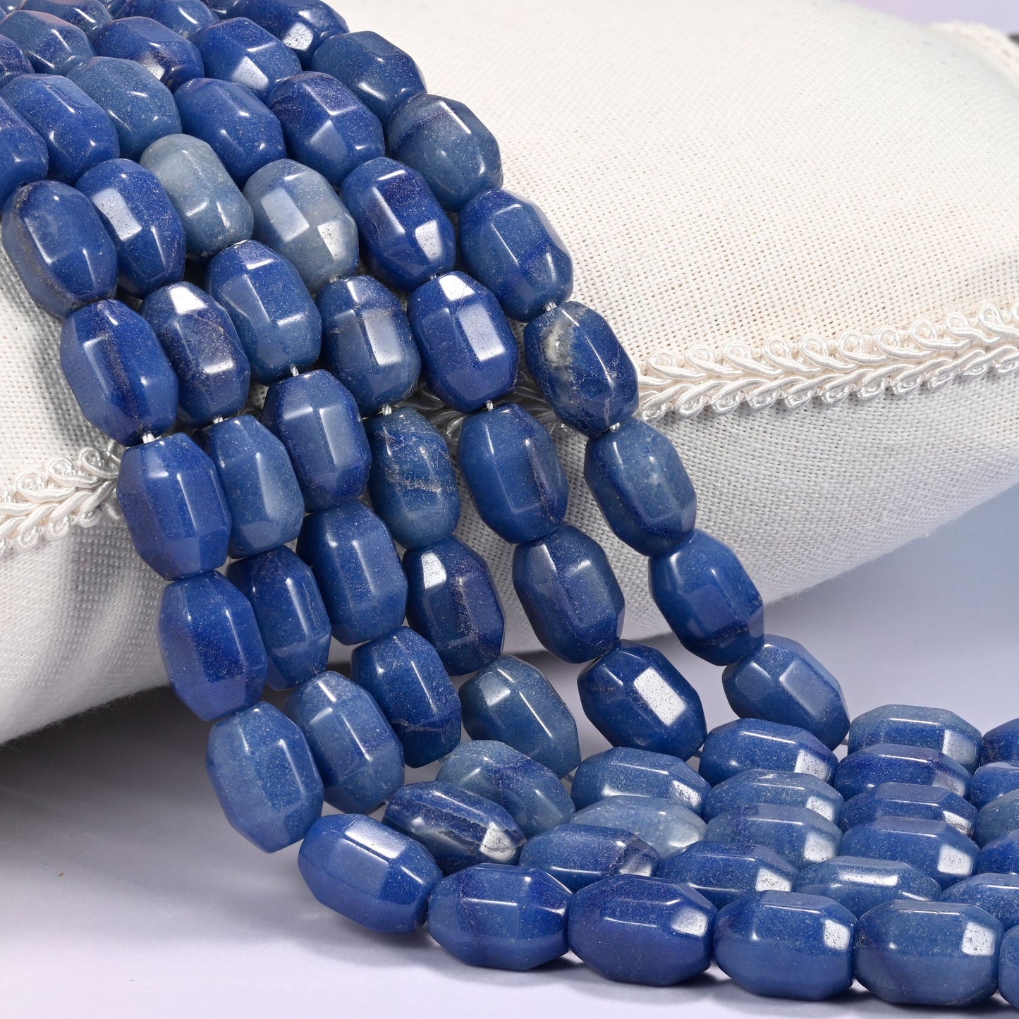 Close-up of multiple strands of deep blue faceted Aventurine barrel beads cascading diagonally over a cream woven pillow with decorative trim.