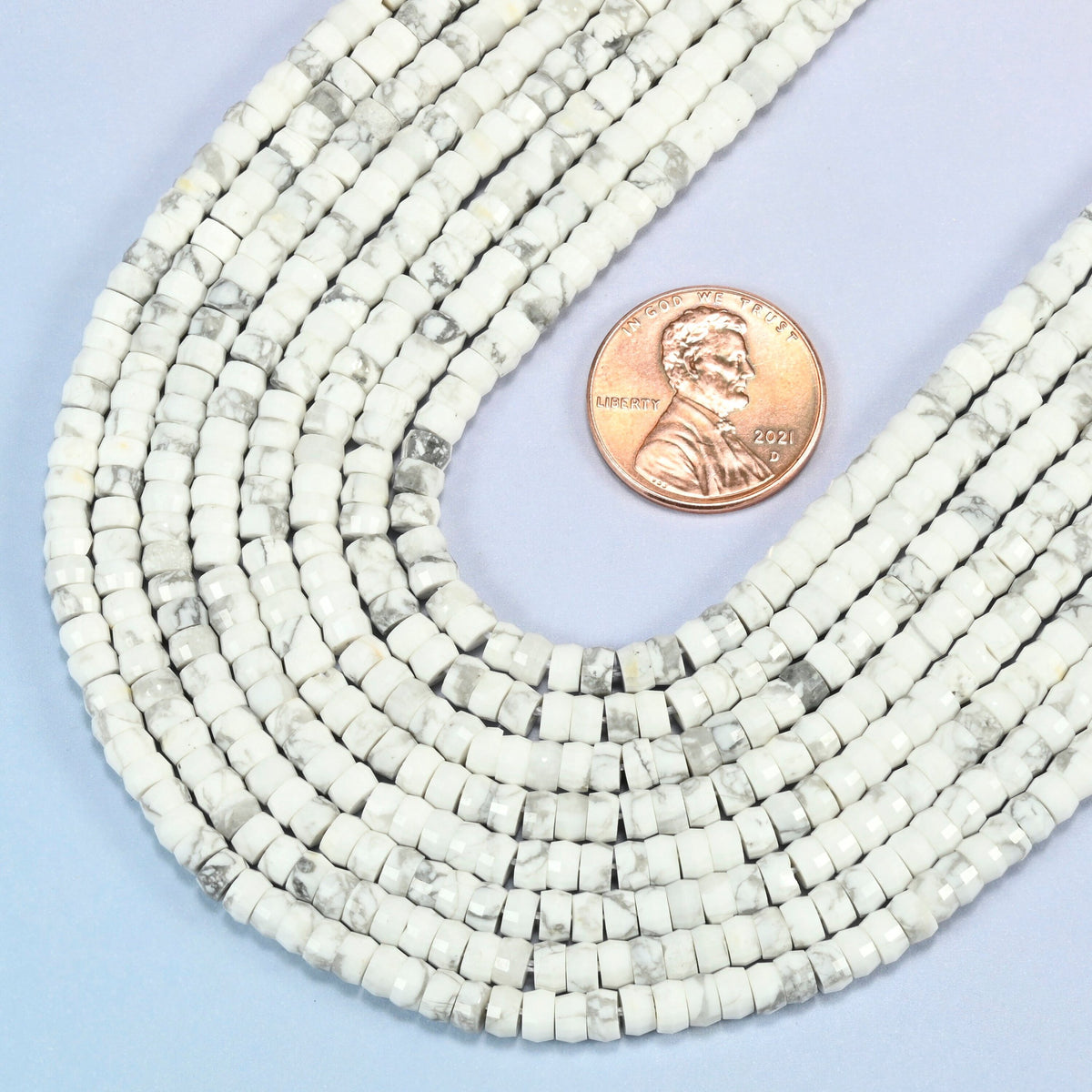 Close view of white howlite faceted heishi disc strands curving beside a US penny for scale