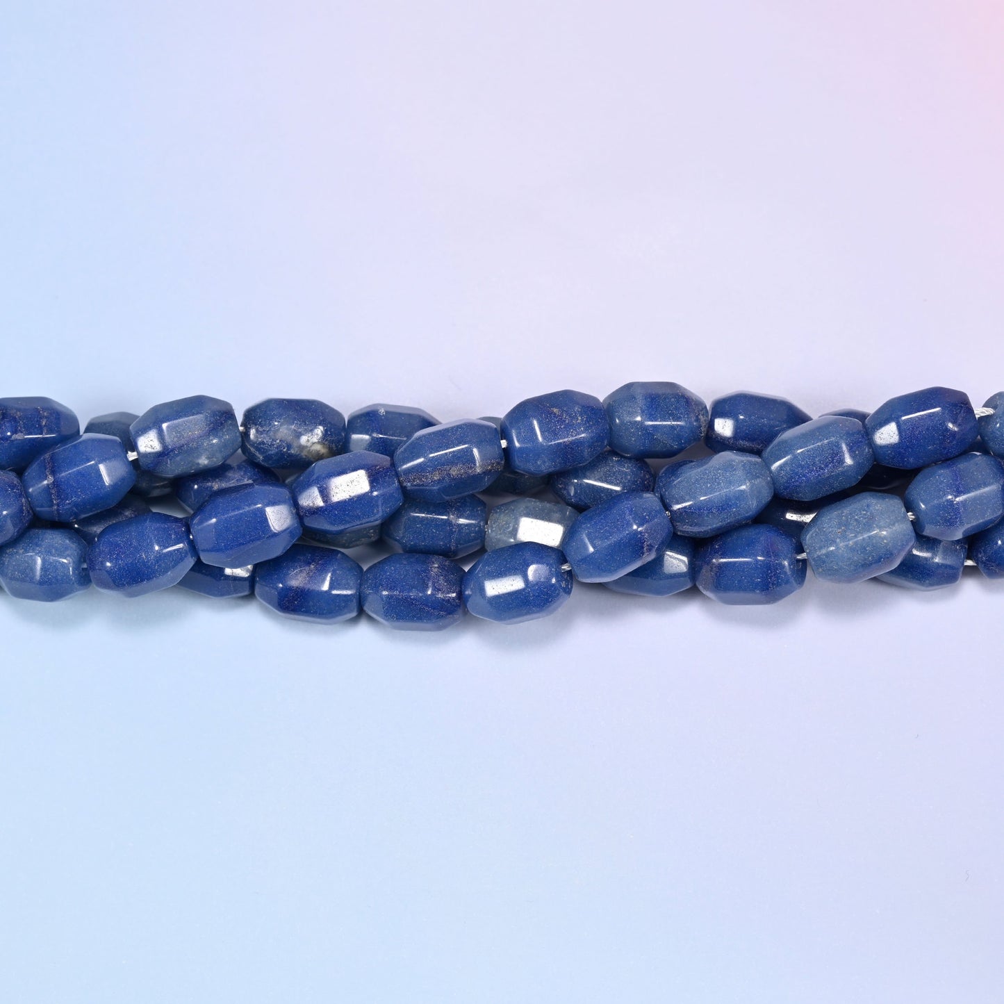 Horizontal row of blue faceted Aventurine barrel beads laid straight across a light blue and lavender gradient background.