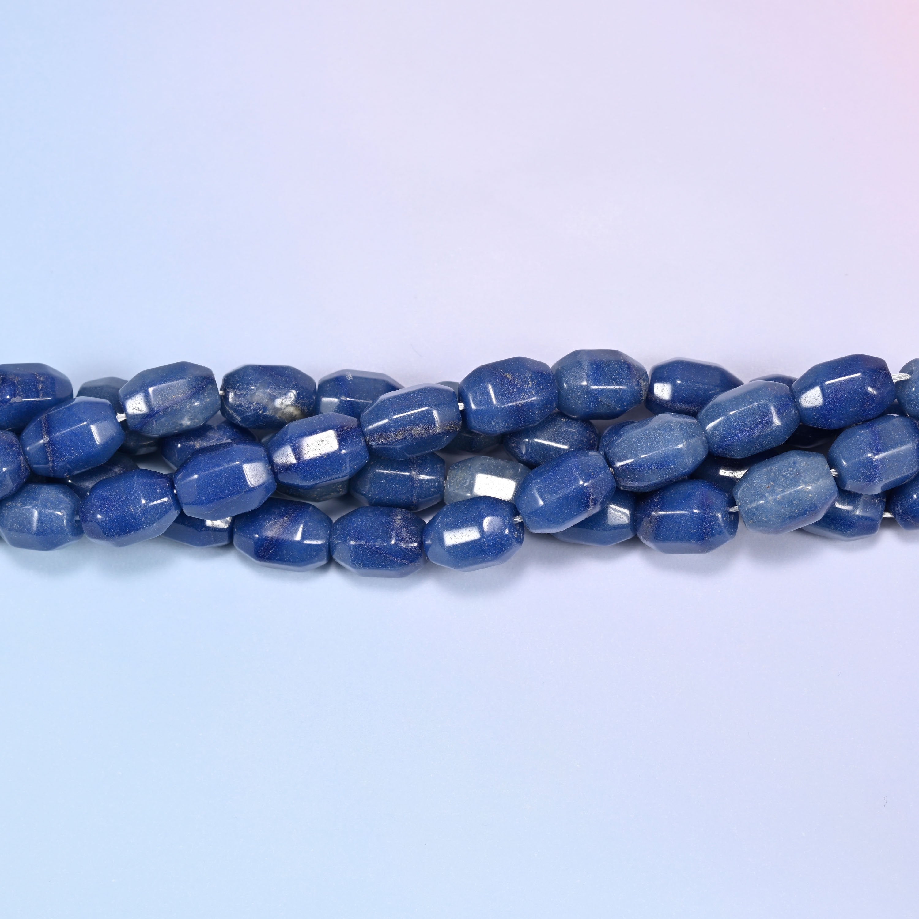 Horizontal row of blue faceted Aventurine barrel beads laid straight across a light blue and lavender gradient background.