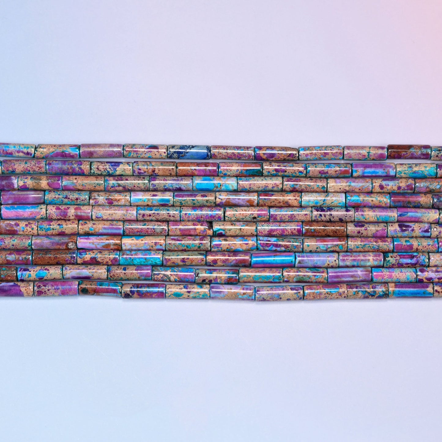 Overhead view of several rows of purple, blue, and turquoise jasper tube beads on a light lavender surface, showing the varied stone patterns.