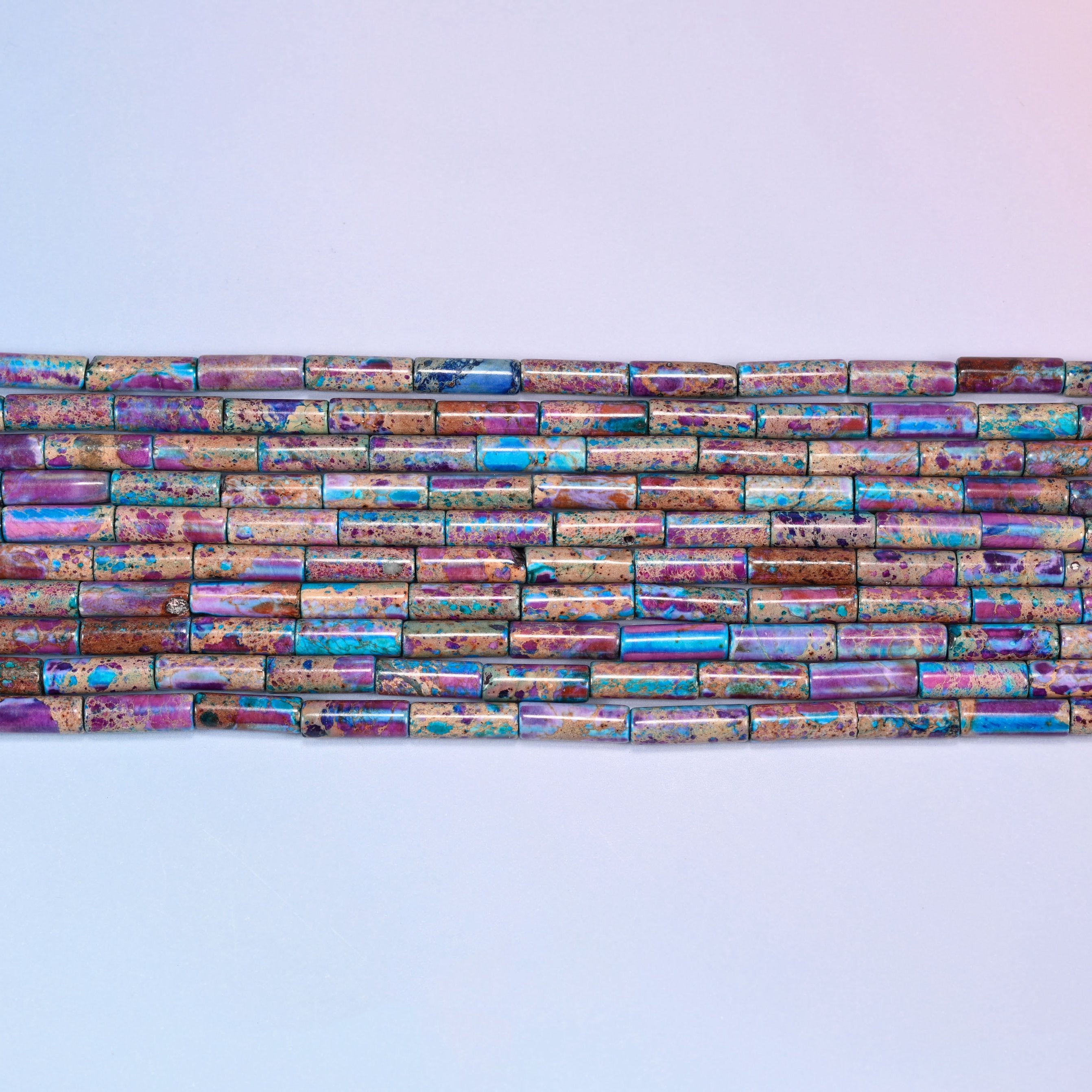 Overhead view of several rows of purple, blue, and turquoise jasper tube beads on a light lavender surface, showing the varied stone patterns.