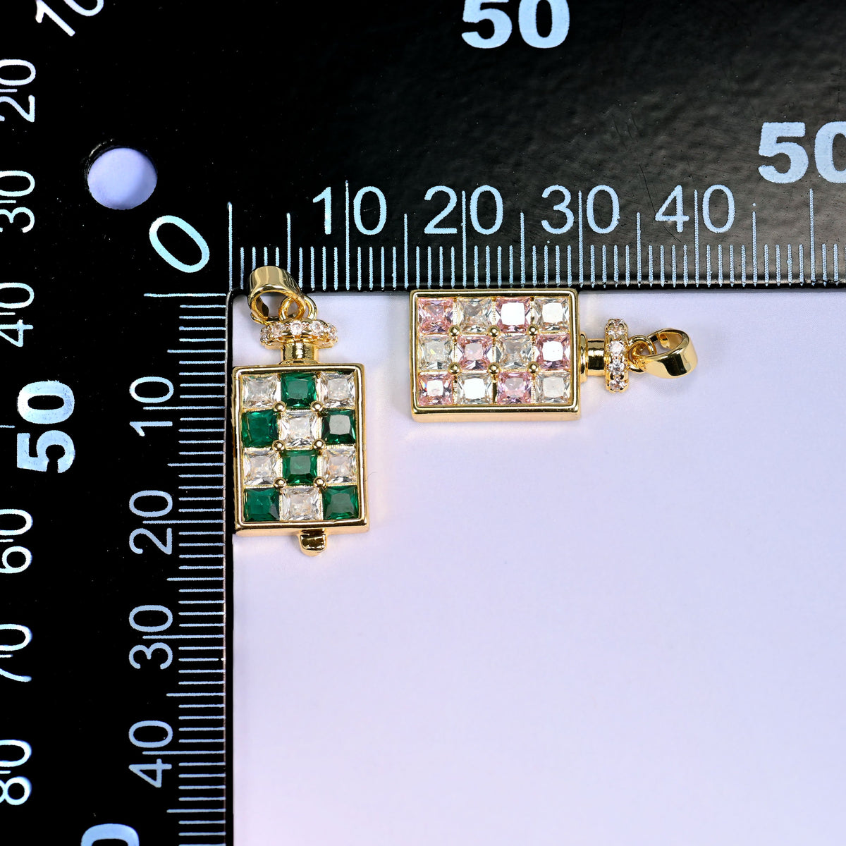 Two perfume bottle zirconia charms on a millimetre ruler, the green one upright at the zero mark and the pink one laid flat to show height and width.