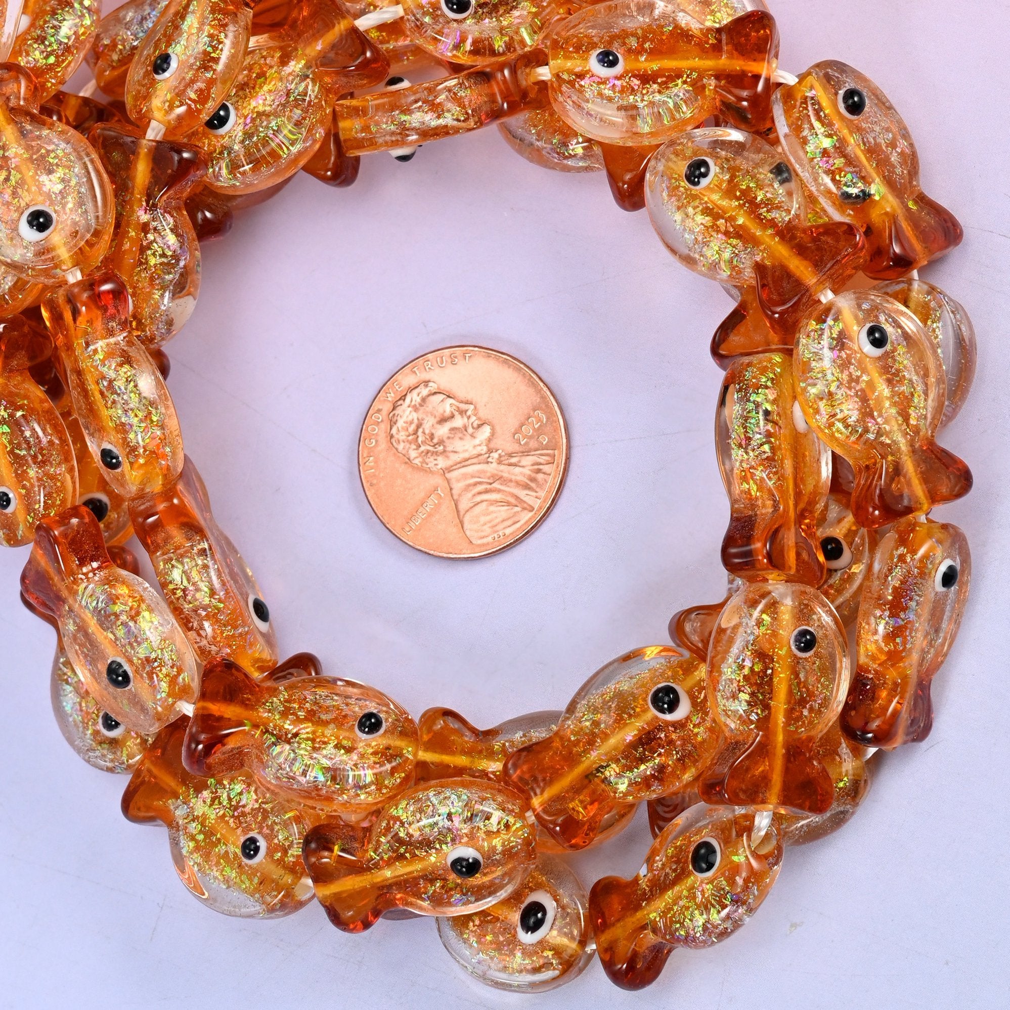 Fish glass beads, 19x12mm, warm amber orange with a clear body dusted with gold glitter and a deeper caramel wash across the head and fanned tail.
