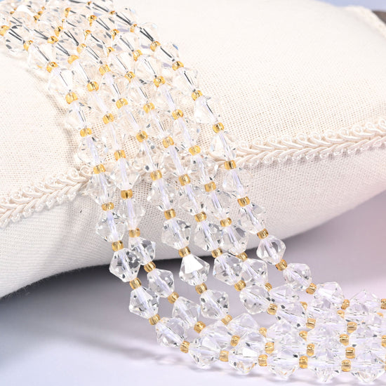 Clear Quartz faceted bicone beads, 8mm, draped diagonally over a white fabric cushion with gold-tone spacer beads between each bead.