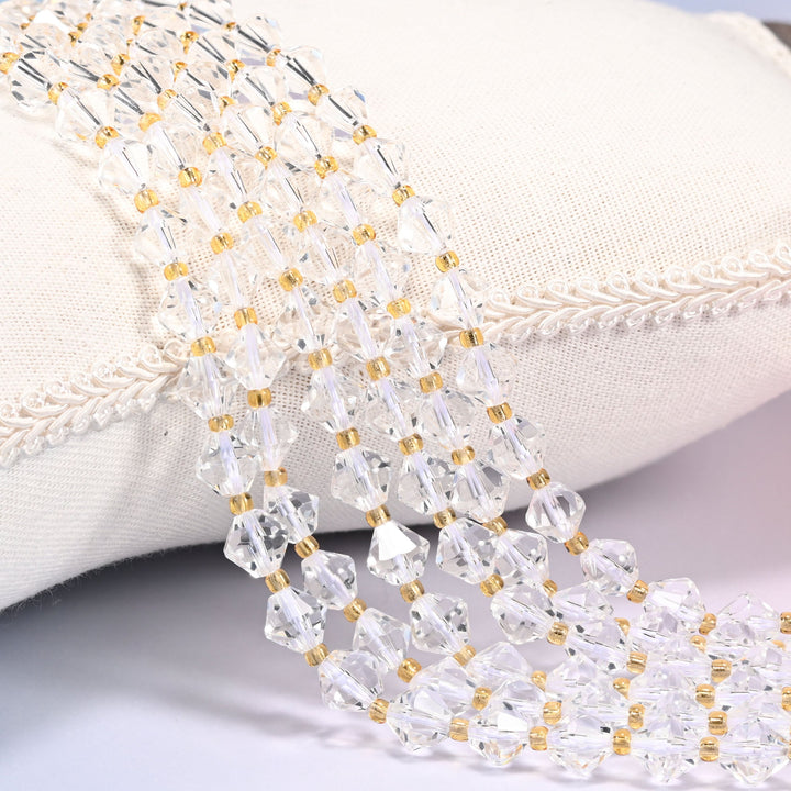 Clear Quartz faceted bicone beads, 8mm, draped diagonally over a white fabric cushion with gold-tone spacer beads between each bead.