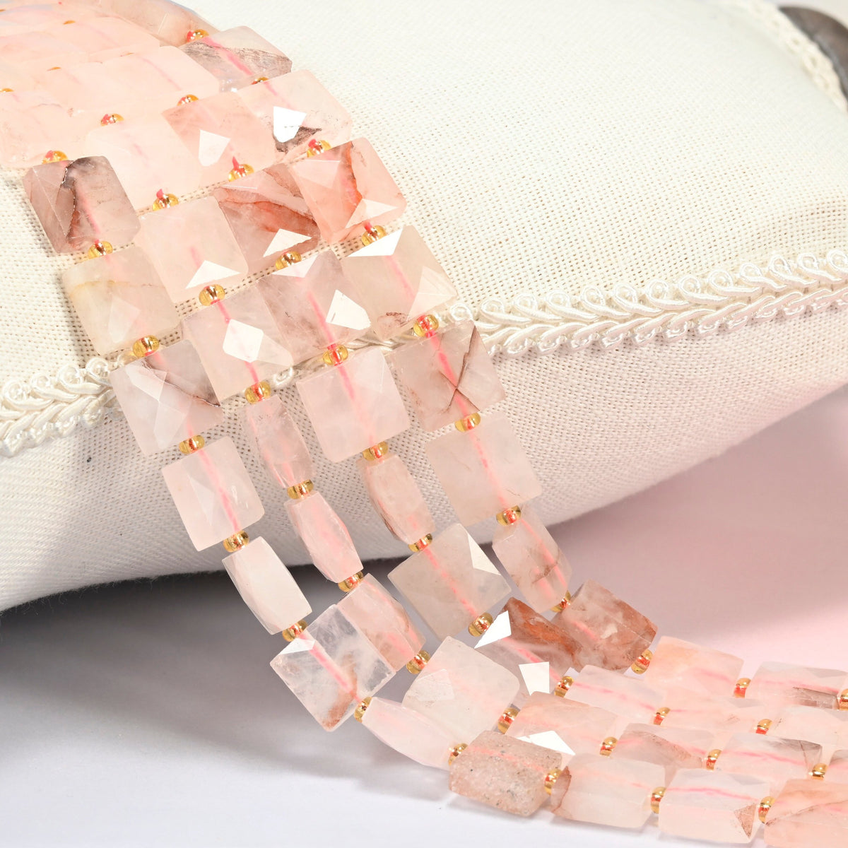 Natural red hematoid quartz faceted flat square beads 10x10mm with small gold seed bead spacers, several strands draped over a white cushion