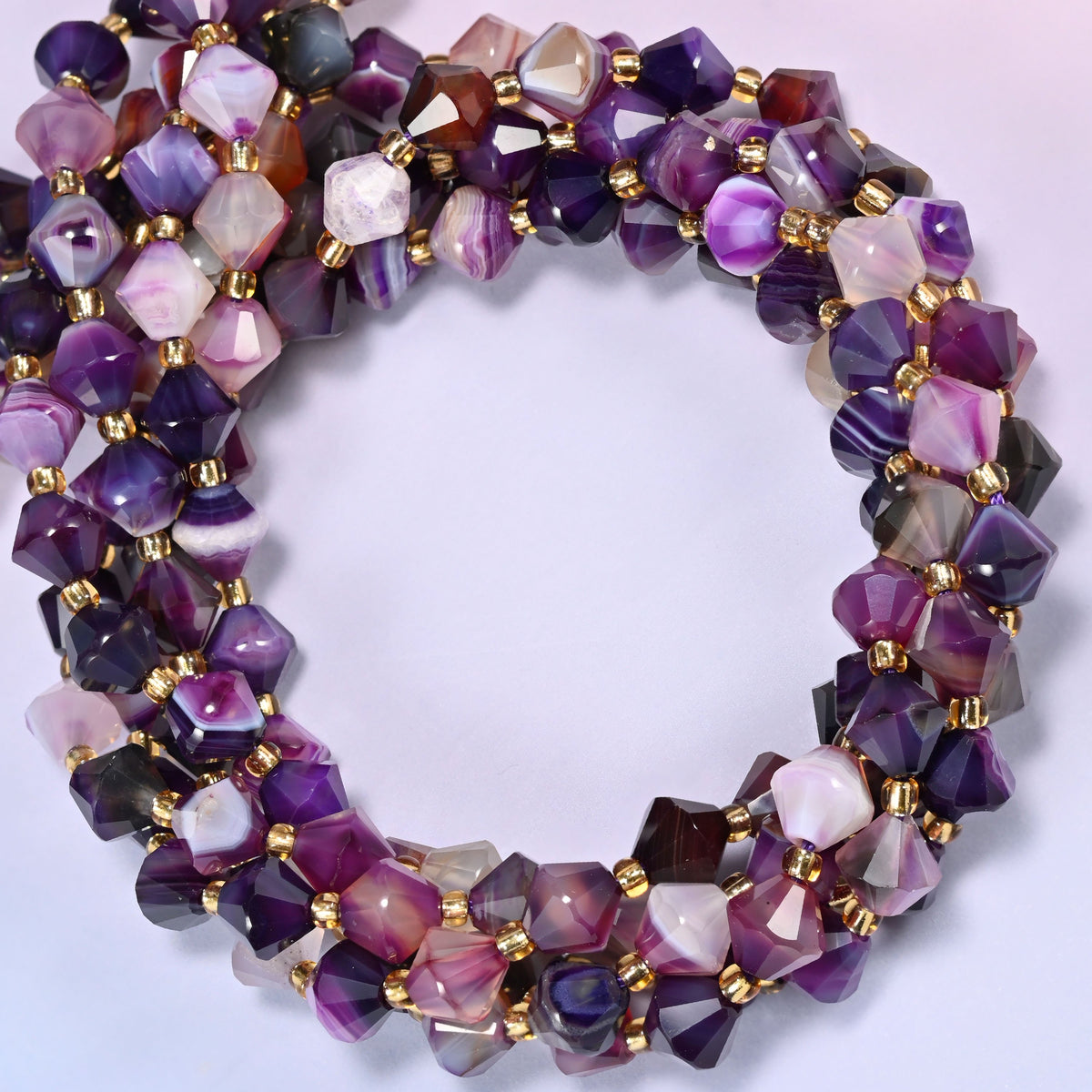 8mm Dyed Purple Strip Agate faceted bicone beads coiled into a circle on a light lavender background, showing shades of purple and pink.