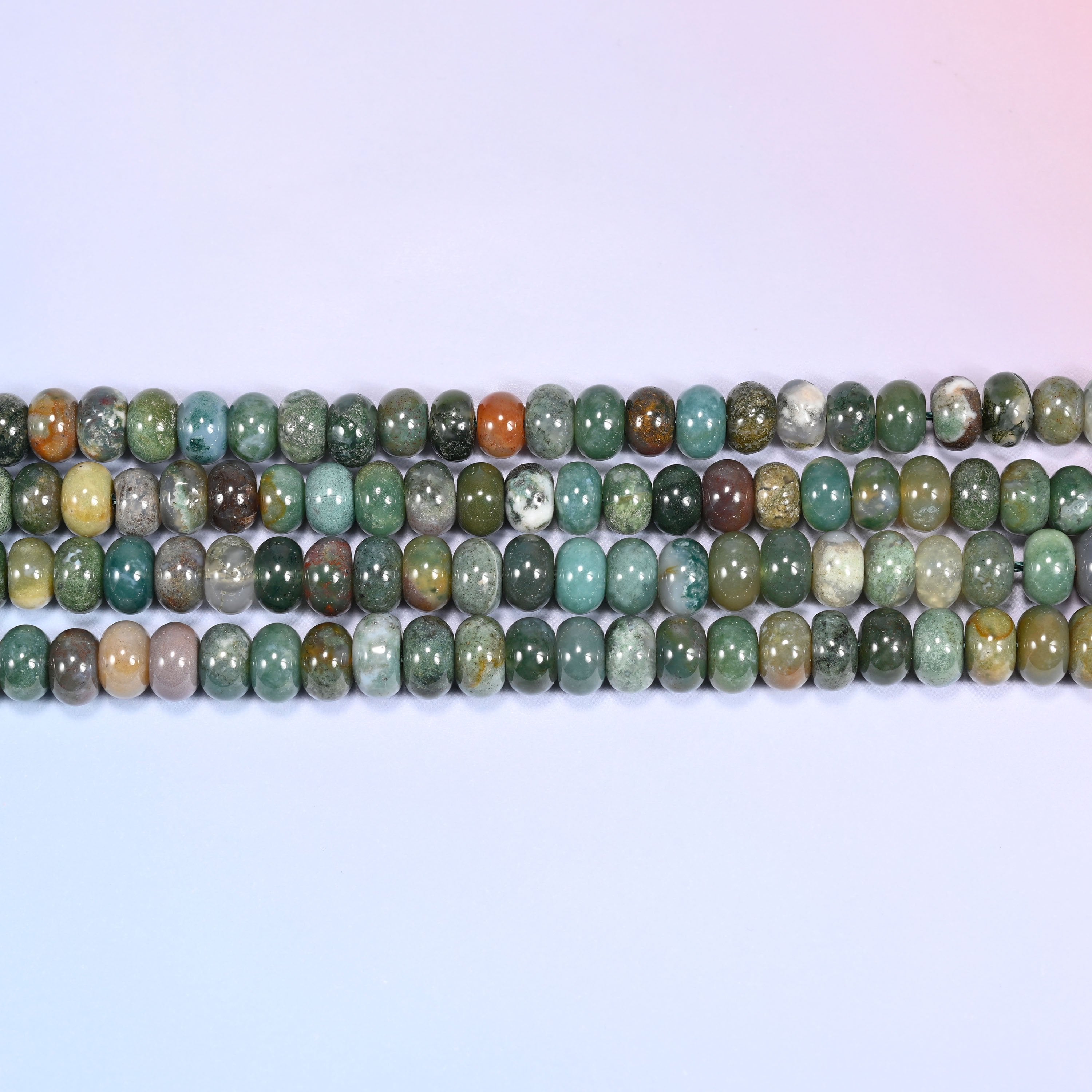 Overhead view of several rows of Indian Agate rondelle beads showing the multicolor green and rust patterning on a light lavender surface.