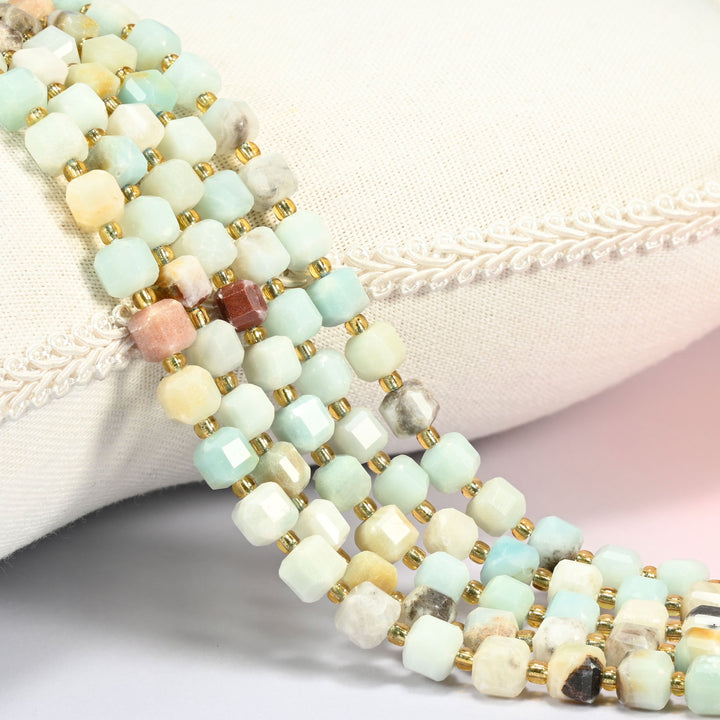 Multi-color amazonite faceted cube beads 6mm with small gold spacer beads, several strands draped over a white cushion
