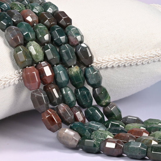 Close-up of multiple strands of dark green and rust red faceted Indian Agate barrel beads cascading diagonally over a cream woven pillow with decorative trim.