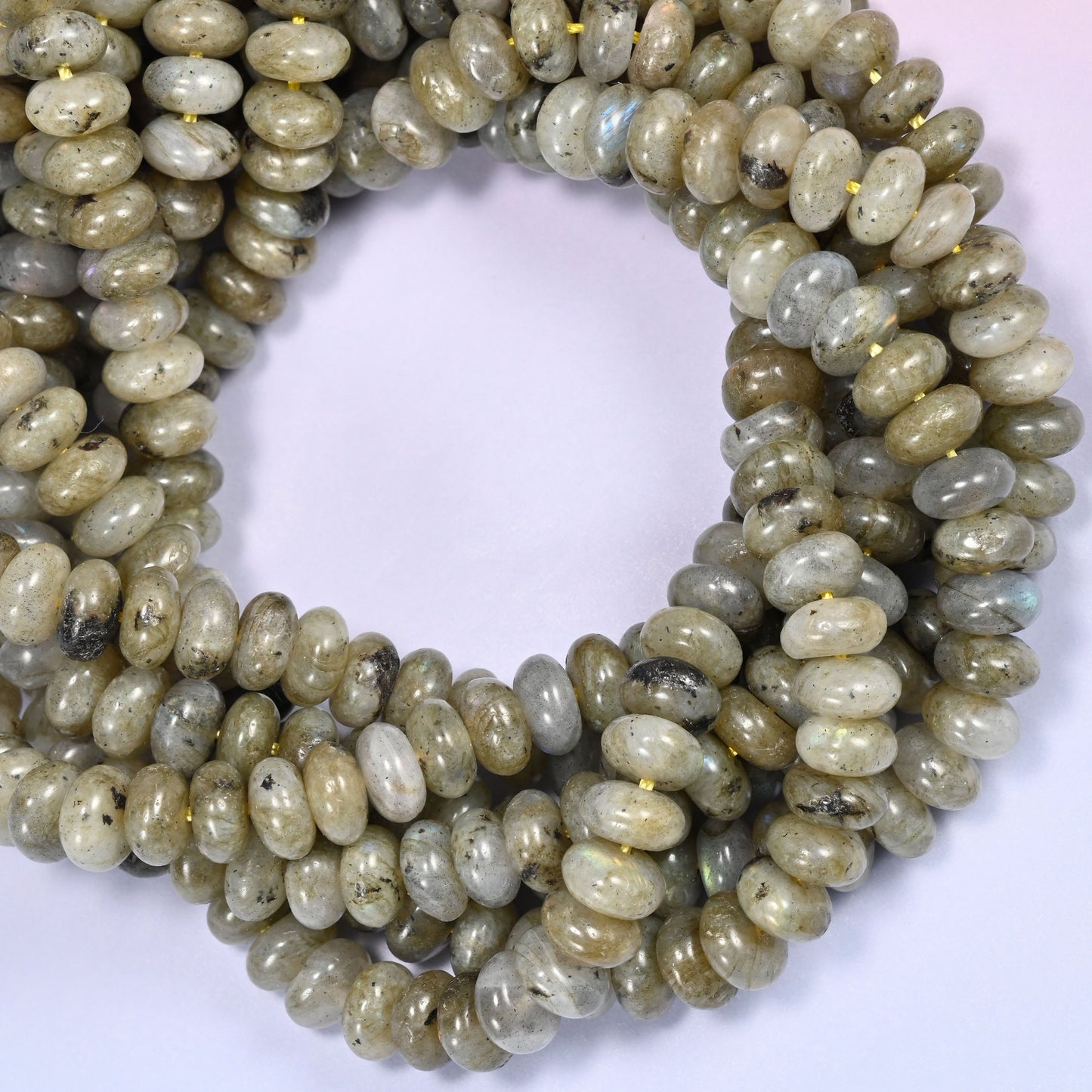 Multiple strands of Grey Labradorite beads gathered together on a lavender surface, loose ends visible.