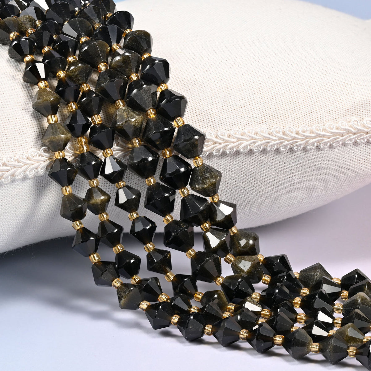 Gold obsidian bicone beads laid out on the diagonal in several rows, dark volcanic glass carrying a warm golden sheen, a gold-tone spacer between every bead.