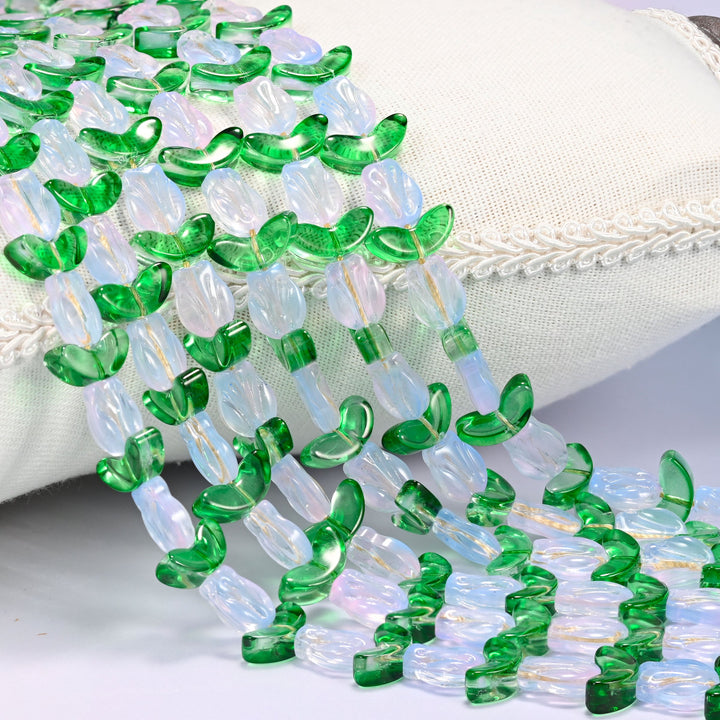 Close-up of white opal-toned tulip flower and green leaf glass beads cascading diagonally over a cream woven pillow with decorative trim.