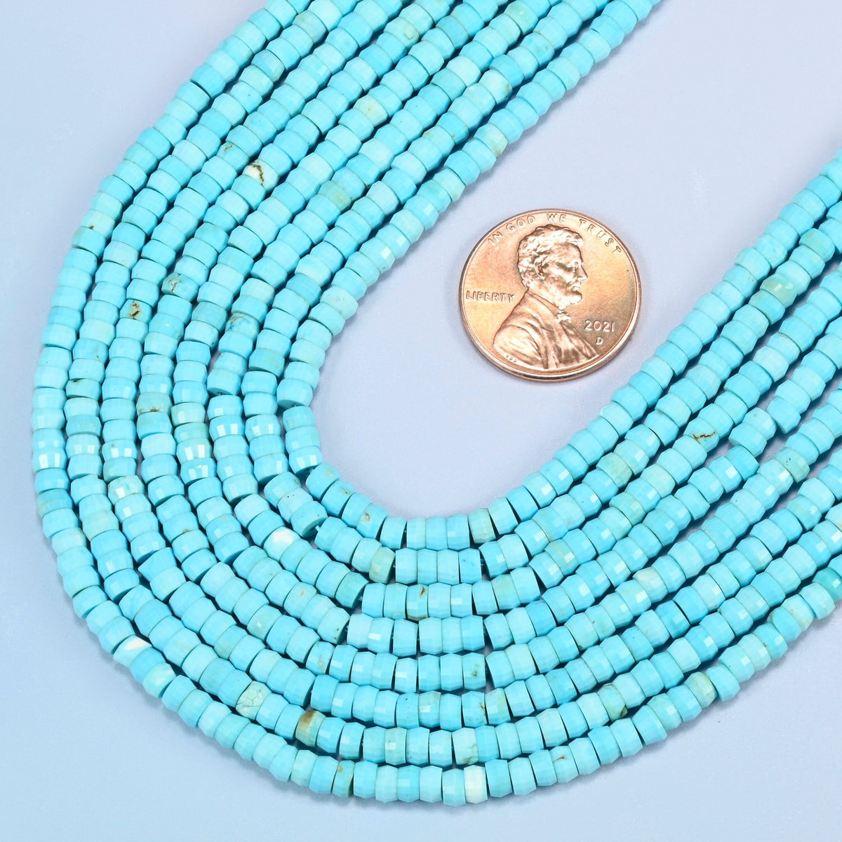 Close view of dyed blue howlite faceted heishi disc strands curving beside a US penny for scale