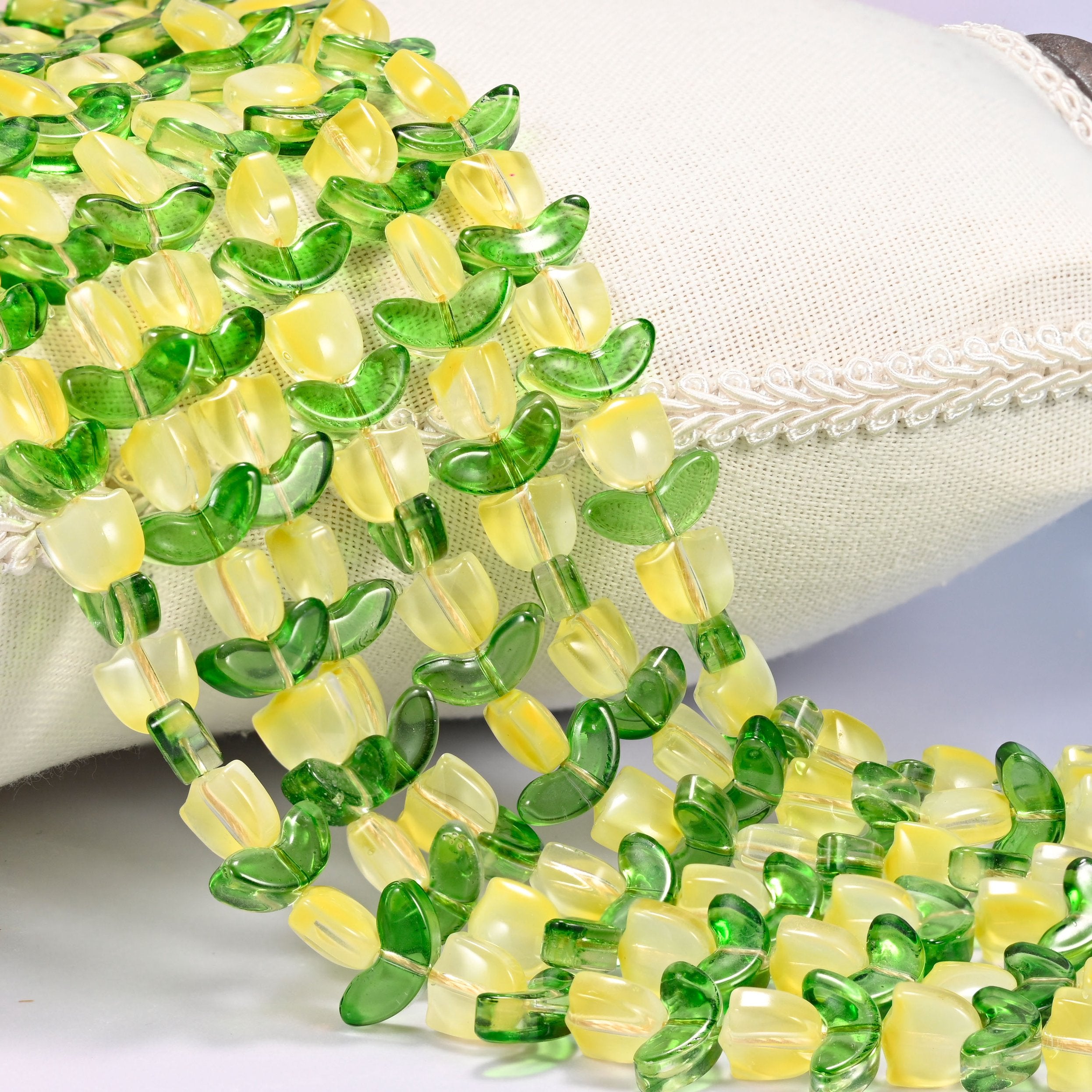 Close-up of yellow tulip flower and green leaf glass beads cascading diagonally over a cream woven pillow with decorative trim.