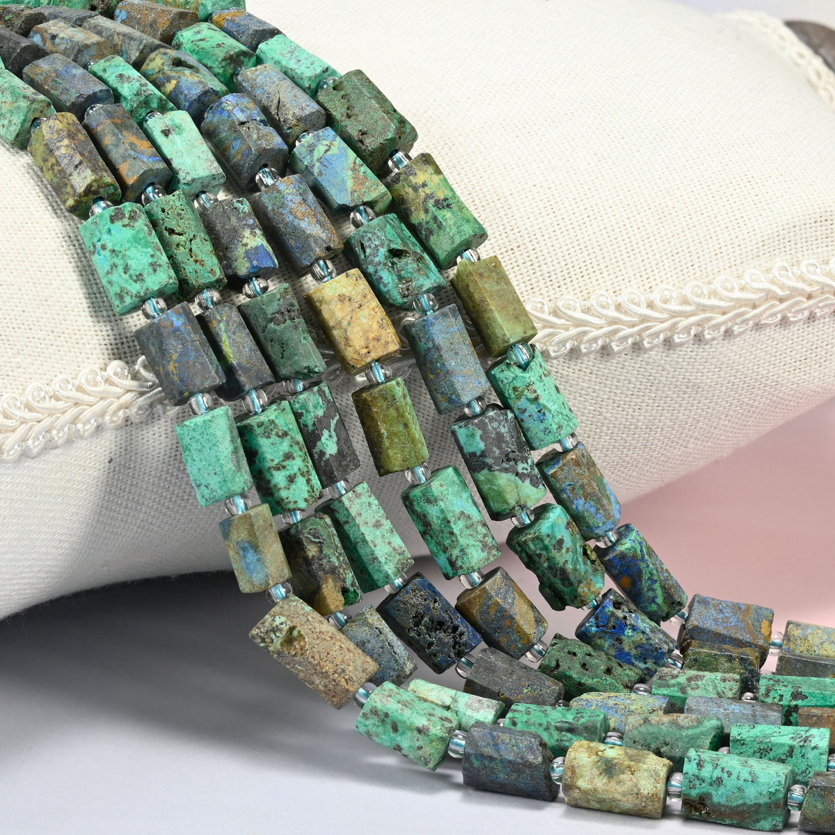 Azurite chrysocolla faceted tube beads 9x7-12x8mm with small clear spacers, several strands draped over a white cushion