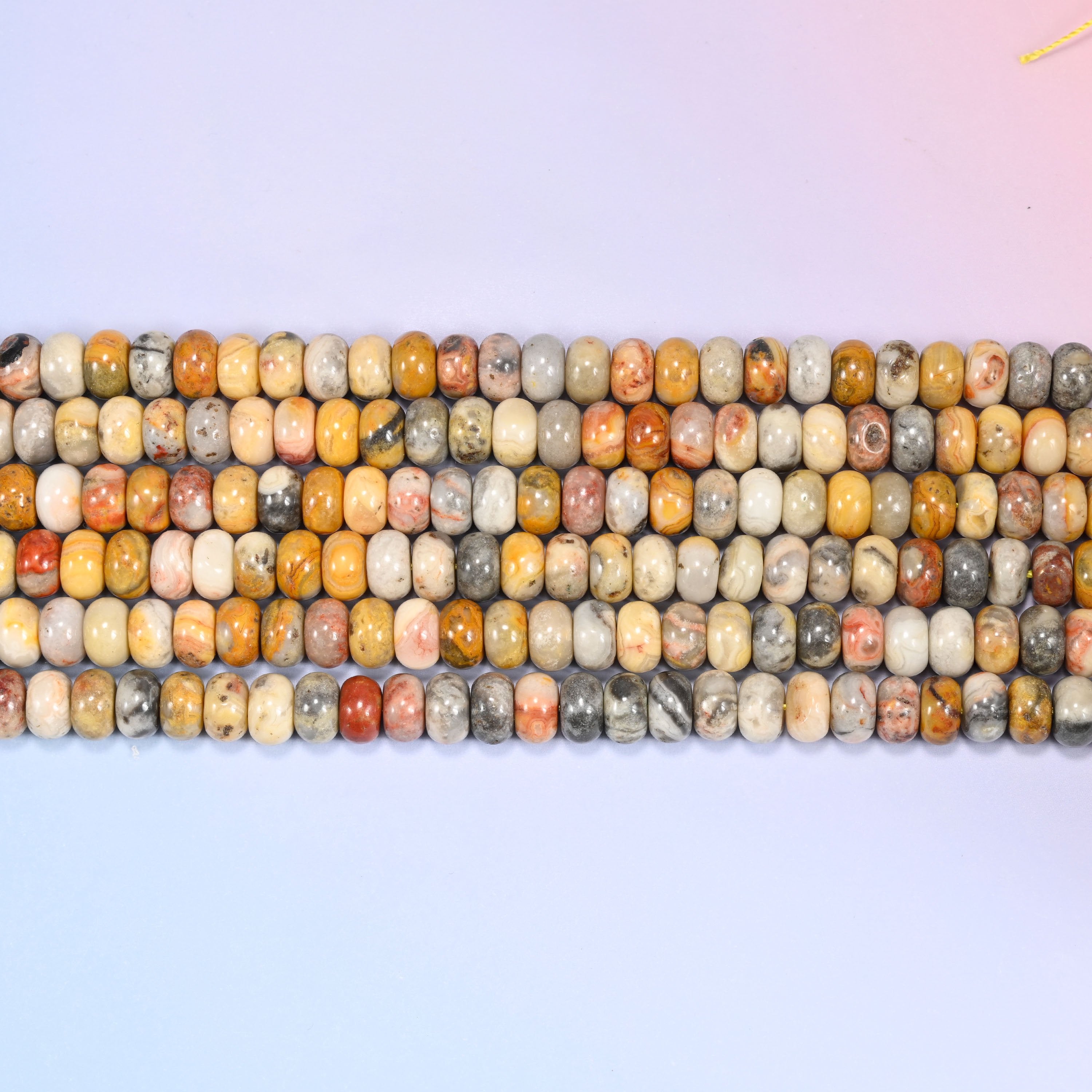 Overhead view of several rows of Crazy Agate rondelle beads showing the mustard, gray, and red veining on a light lavender surface.
