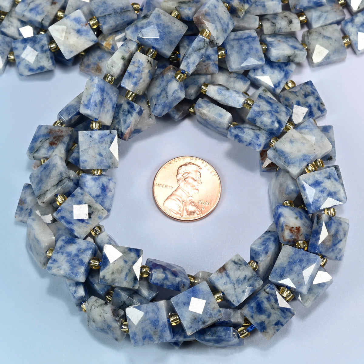 Natural blue spot jasper faceted flat square beads 10x10mm coiled in a loop around a US penny for scale, close-up of the faceted flat square shape