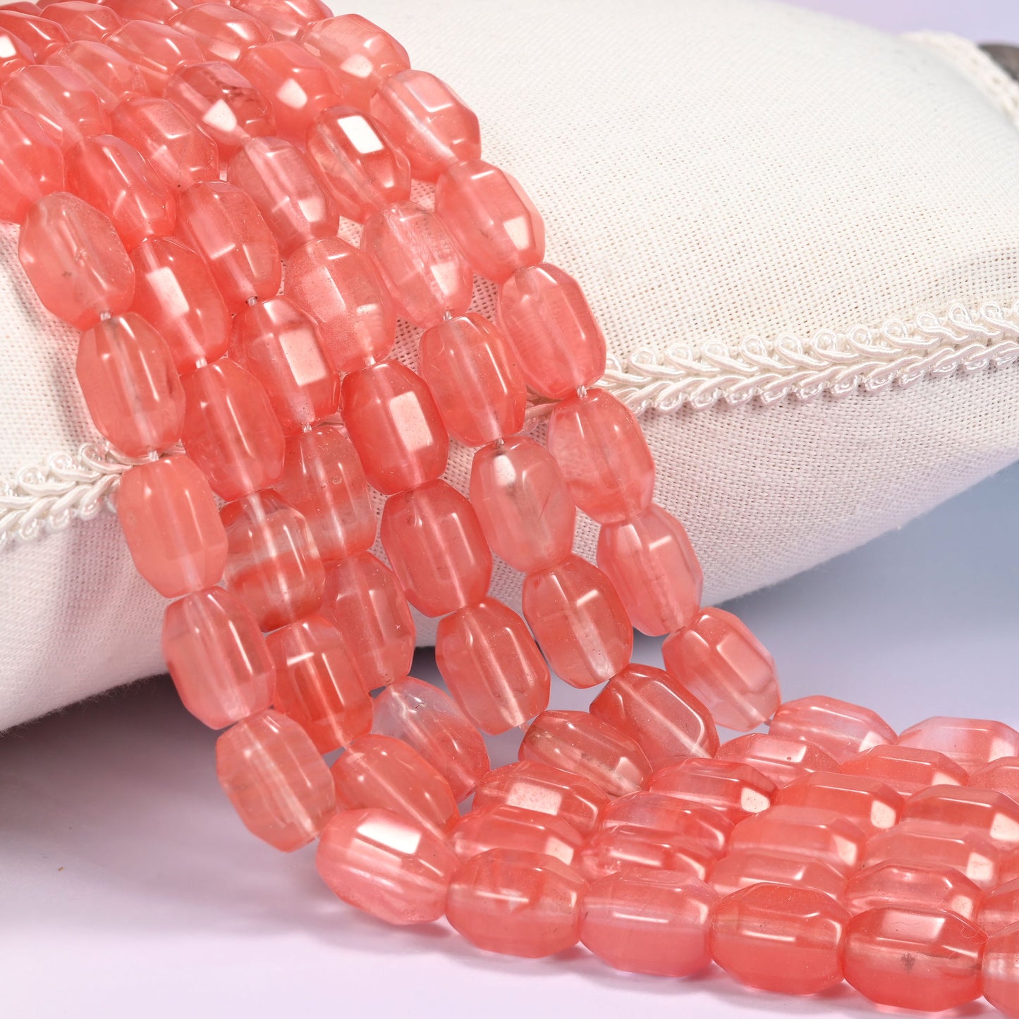 Close-up of multiple strands of coral pink faceted glass barrel beads cascading diagonally over a cream woven pillow with decorative trim.