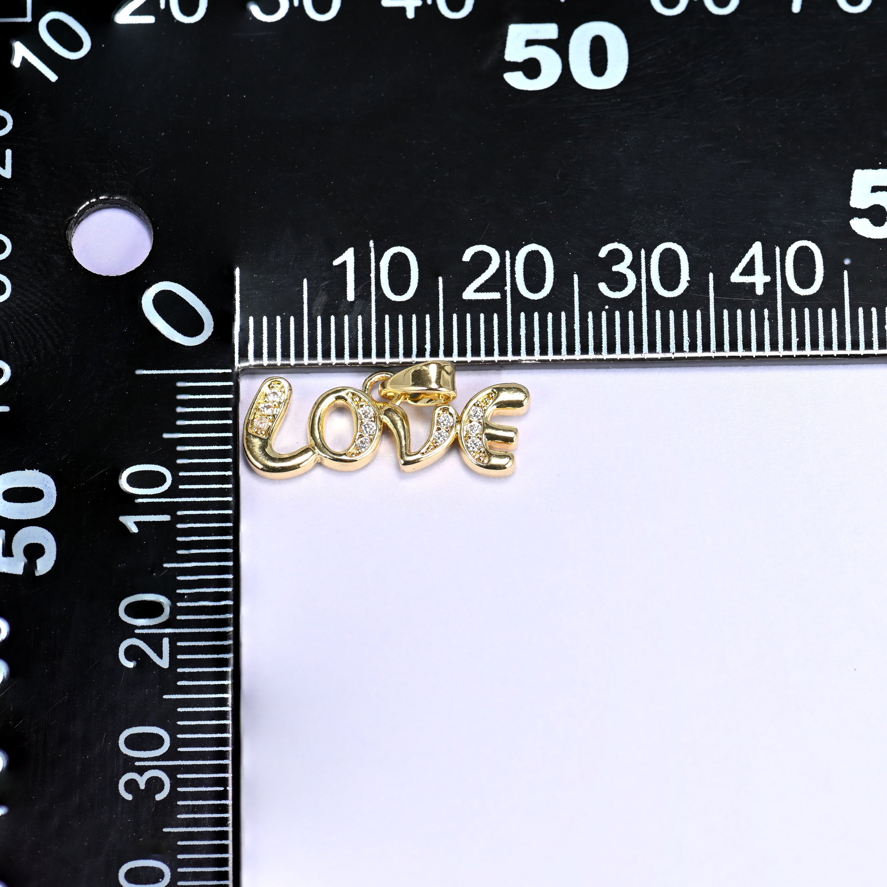 Love letter zirconia charm measured against a black millimetre ruler, the gold Love script pendant spanning roughly 20mm.