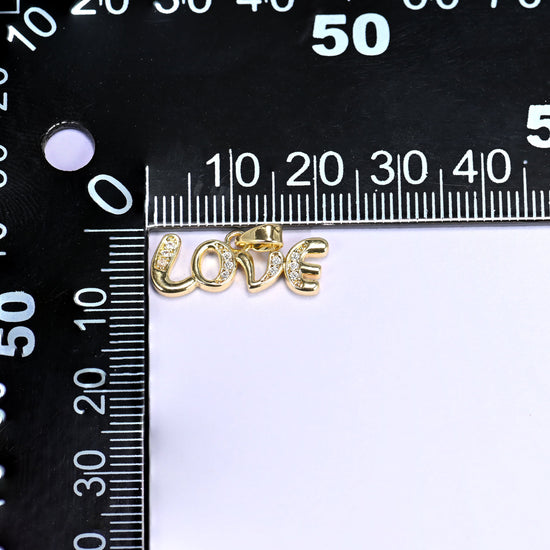 Love letter zirconia charm measured against a black millimetre ruler, the gold Love script pendant spanning roughly 20mm.