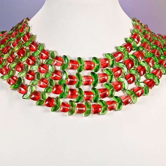 Multi-strand necklace of red tulip and green leaf glass beads laid on a white mannequin neck form against a lavender background.