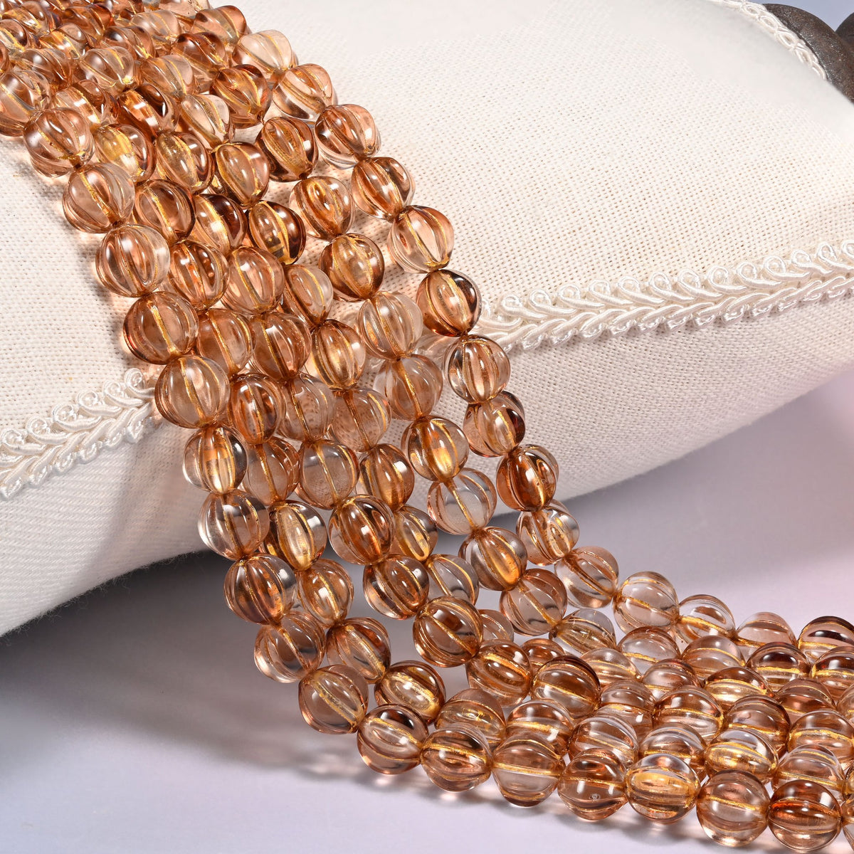 Brown pumpkin glass beads, 8mm fluted melon rounds, several strands draped over a white cushion.