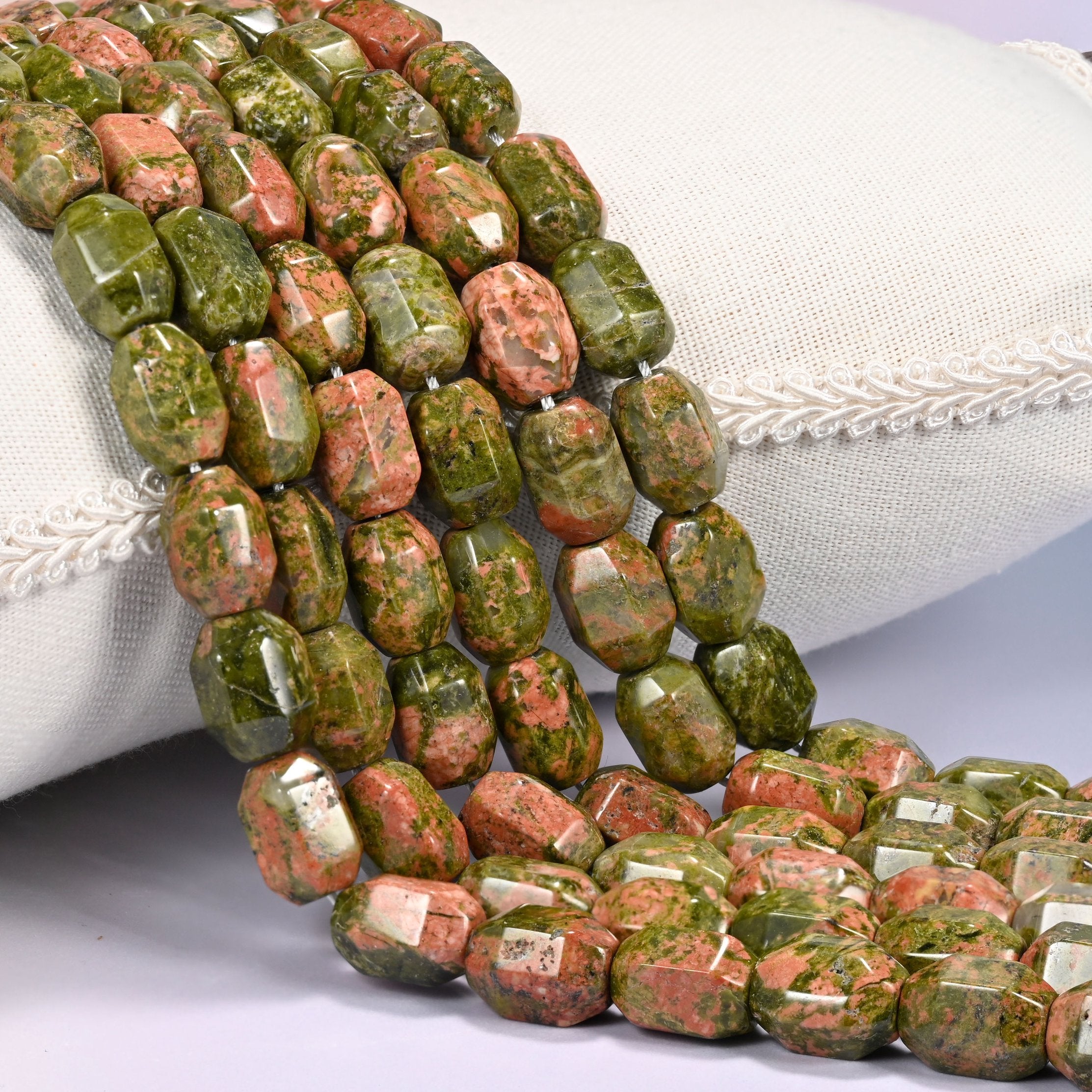 Close-up of multiple strands of green and salmon faceted Unakite barrel beads cascading diagonally over a cream woven pillow with decorative trim.