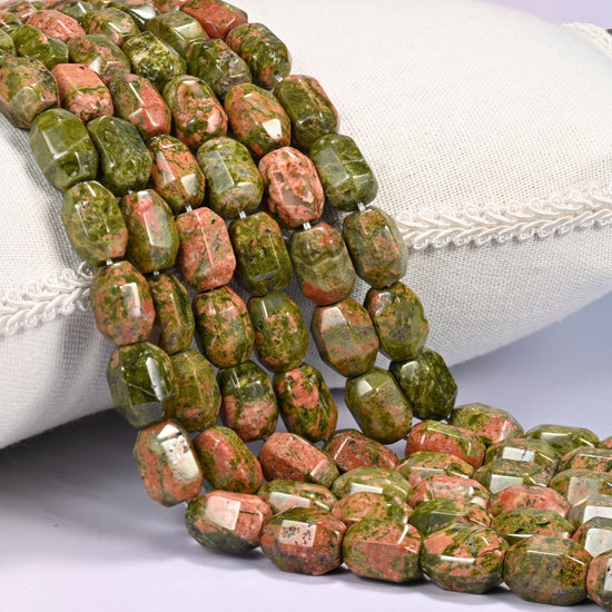 Close-up of multiple strands of green and salmon faceted Unakite barrel beads cascading diagonally over a cream woven pillow with decorative trim.