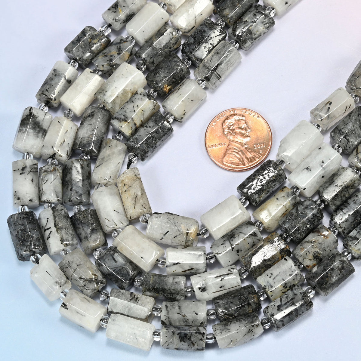 Close view of black rutilated quartz faceted tube strands curving beside a US penny for scale