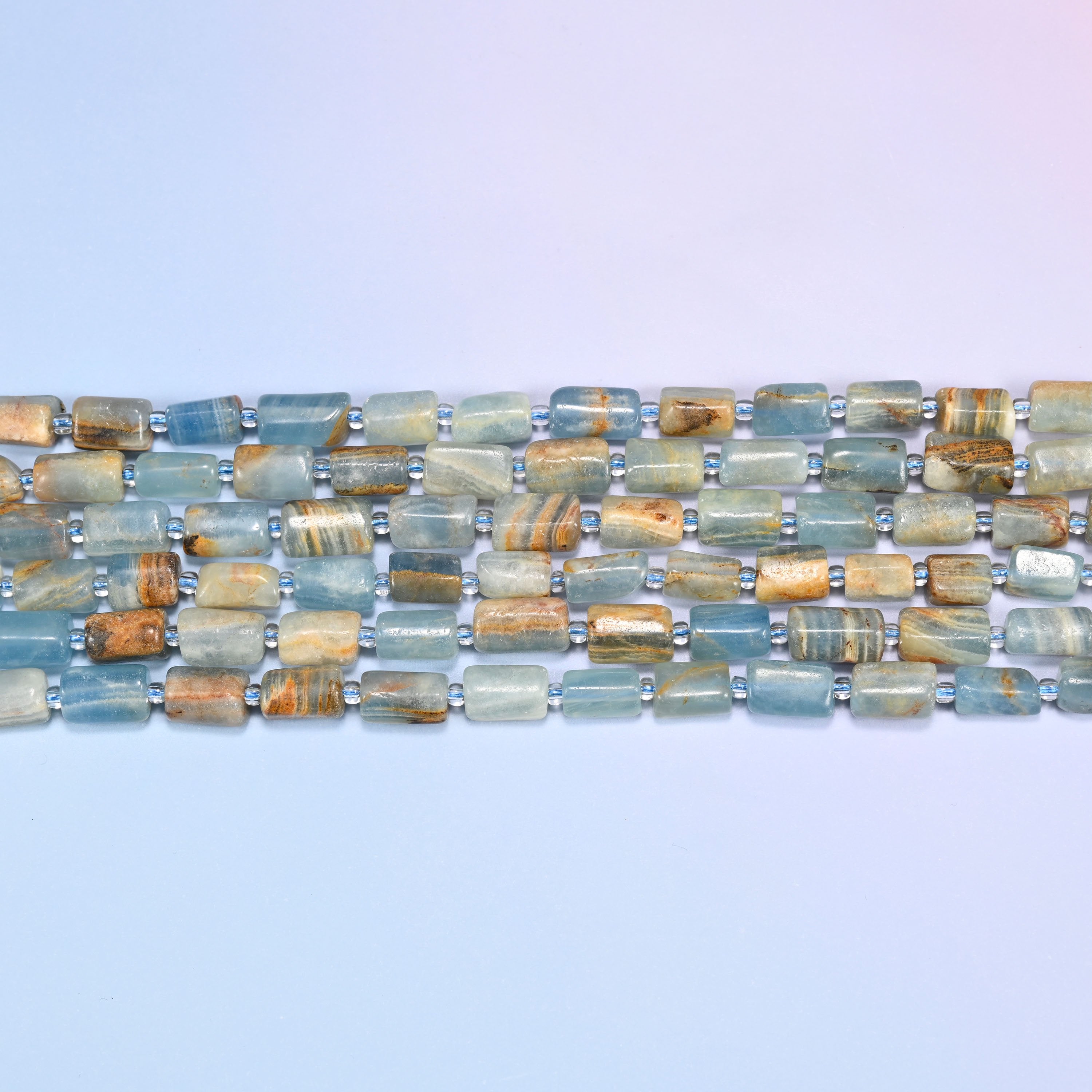 Overhead view of several rows of faceted Blue Calcite tube beads in aqua, tan, and rust striped tones on a light lavender surface.