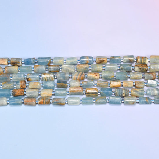 Overhead view of several rows of faceted Blue Calcite tube beads in aqua, tan, and rust striped tones on a light lavender surface.