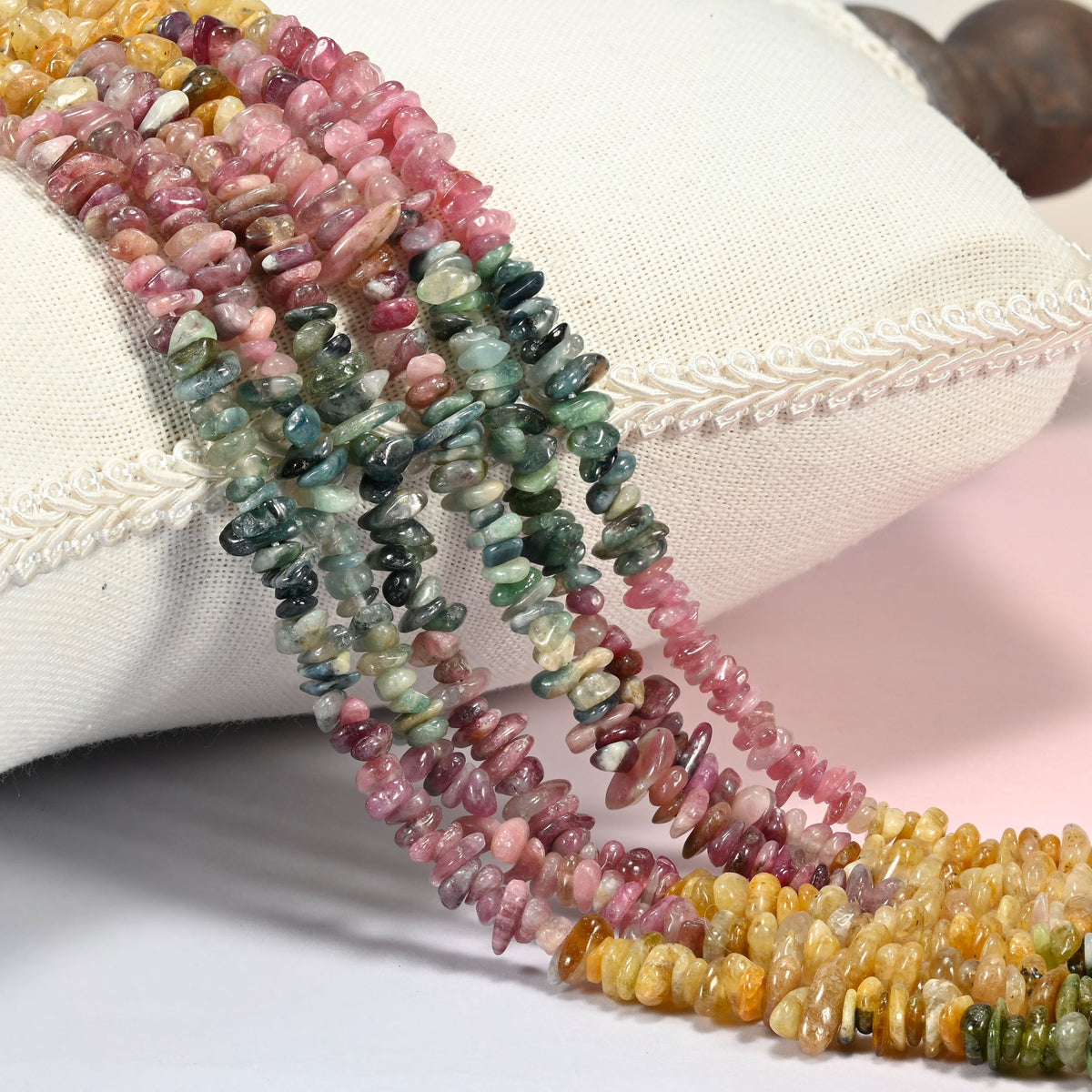 Multicolor tourmaline chip beads 3-8mm, several strands draped over a white cushion in pink, green and golden yellow
