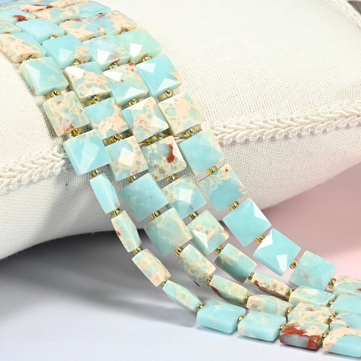 Dyed blue Shoushan stone faceted flat square beads 10x10mm with small gold seed bead spacers, several strands draped over a white cushion
