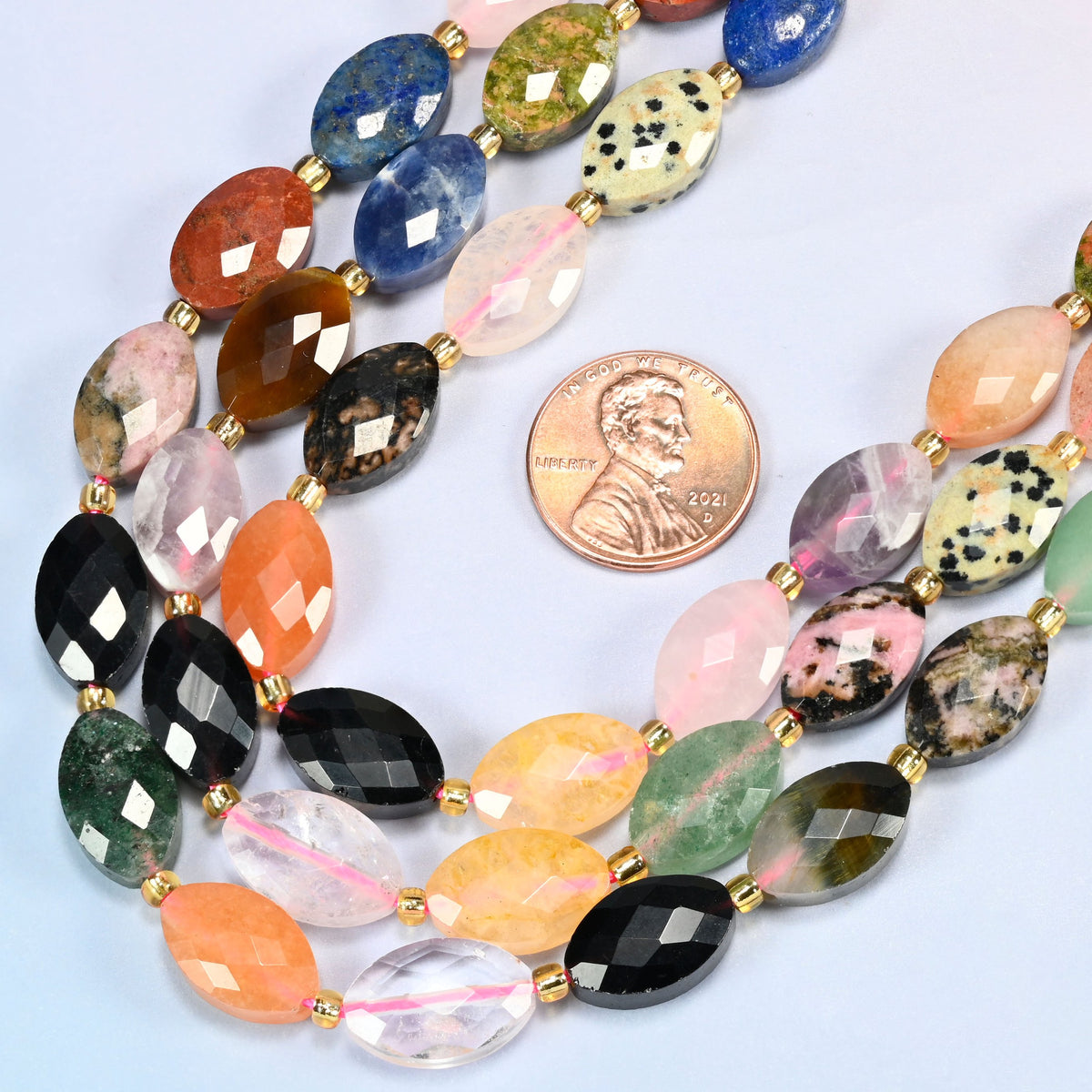 Close view of assorted gemstone faceted flat oval strands curving beside a US penny for scale