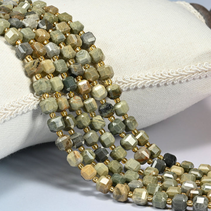 Silver leaf jasper faceted cube beads with gold glass spacers, several strands draped over a white linen cushion.