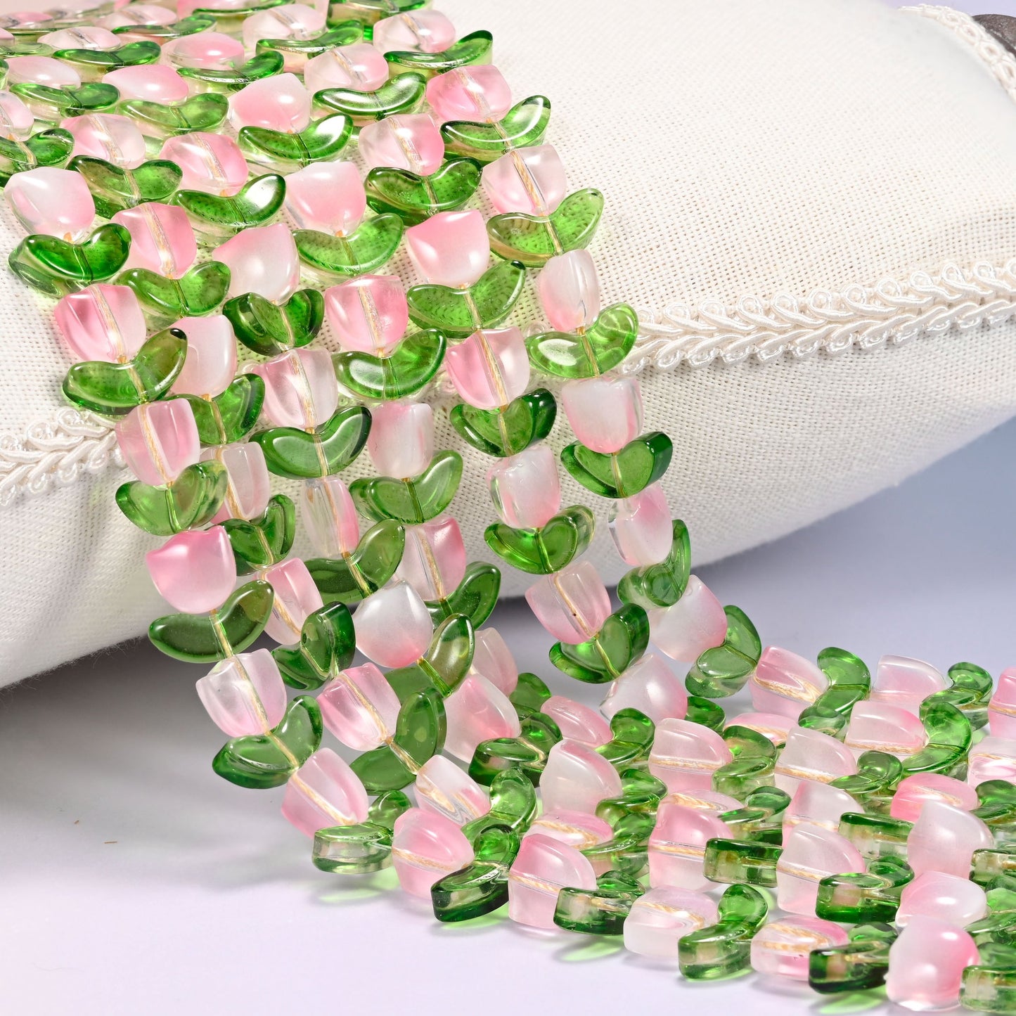 Close-up of pink tulip flower and green leaf glass beads cascading diagonally over a cream woven pillow with decorative trim.