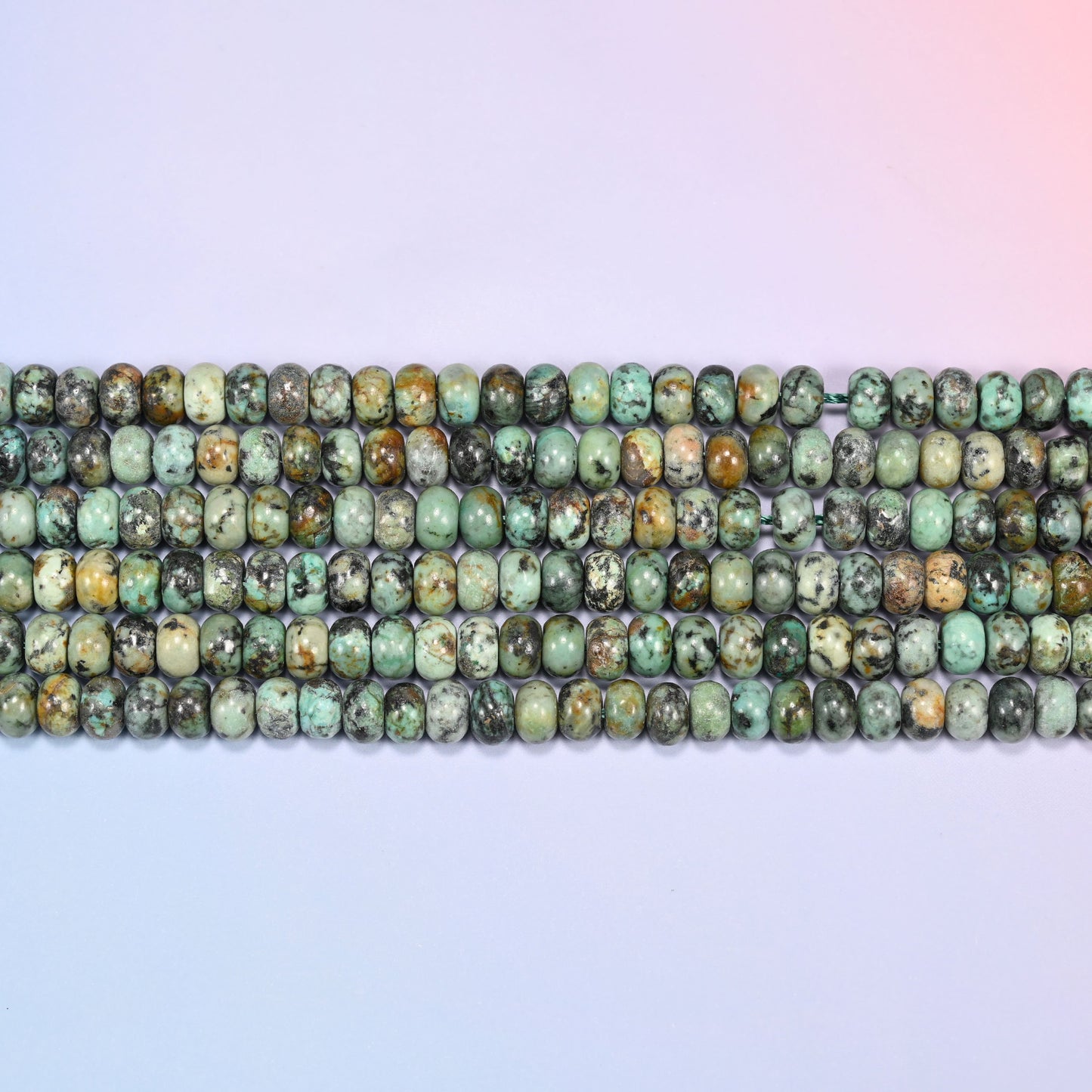 Overhead view of several rows of African Howlite rondelle beads showing the green and dark matrix coloring on a light lavender surface.