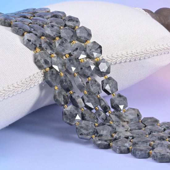 Black Labradorite faceted hexagon strands resting on a white cushion, grey stone flecked with charcoal