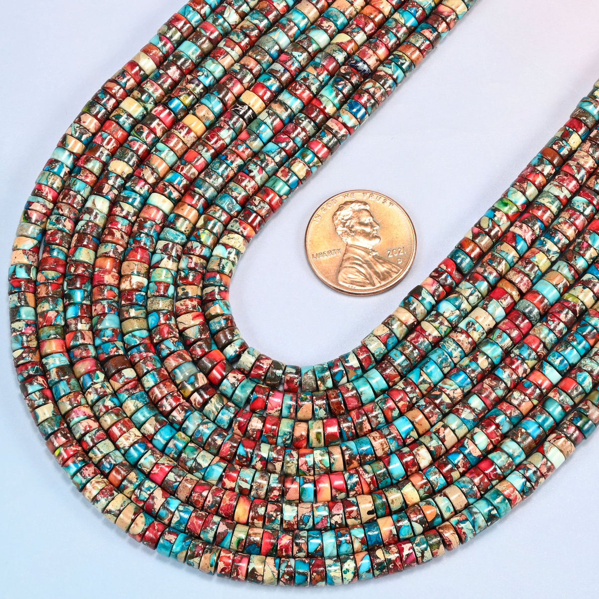 Close view of dyed red blue sea sediment jasper heishi disc strands curving beside a US penny for scale