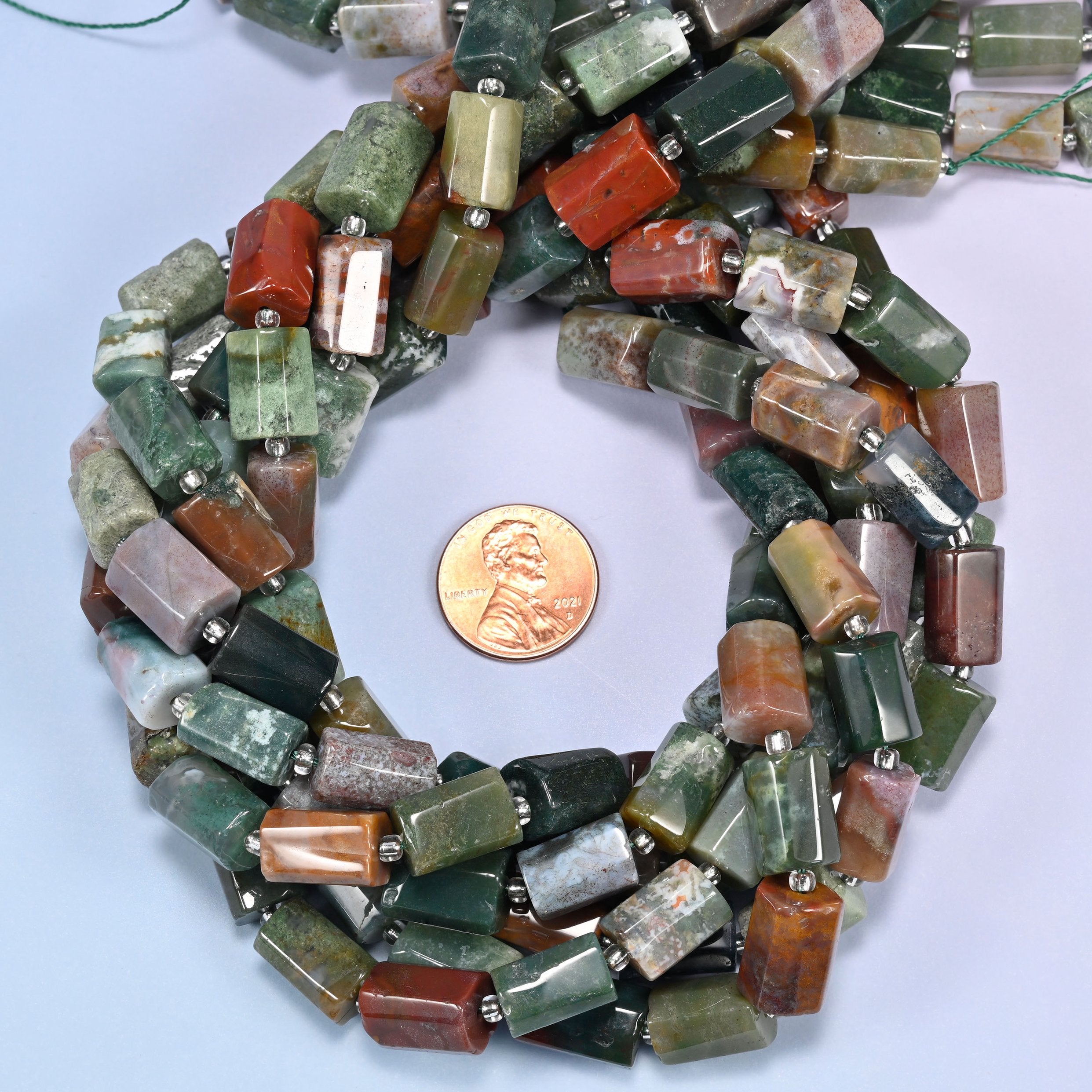 Bundled coil of faceted Indian Agate tube beads with clear glass spacer beads on a lavender background with a penny placed at the center for scale.