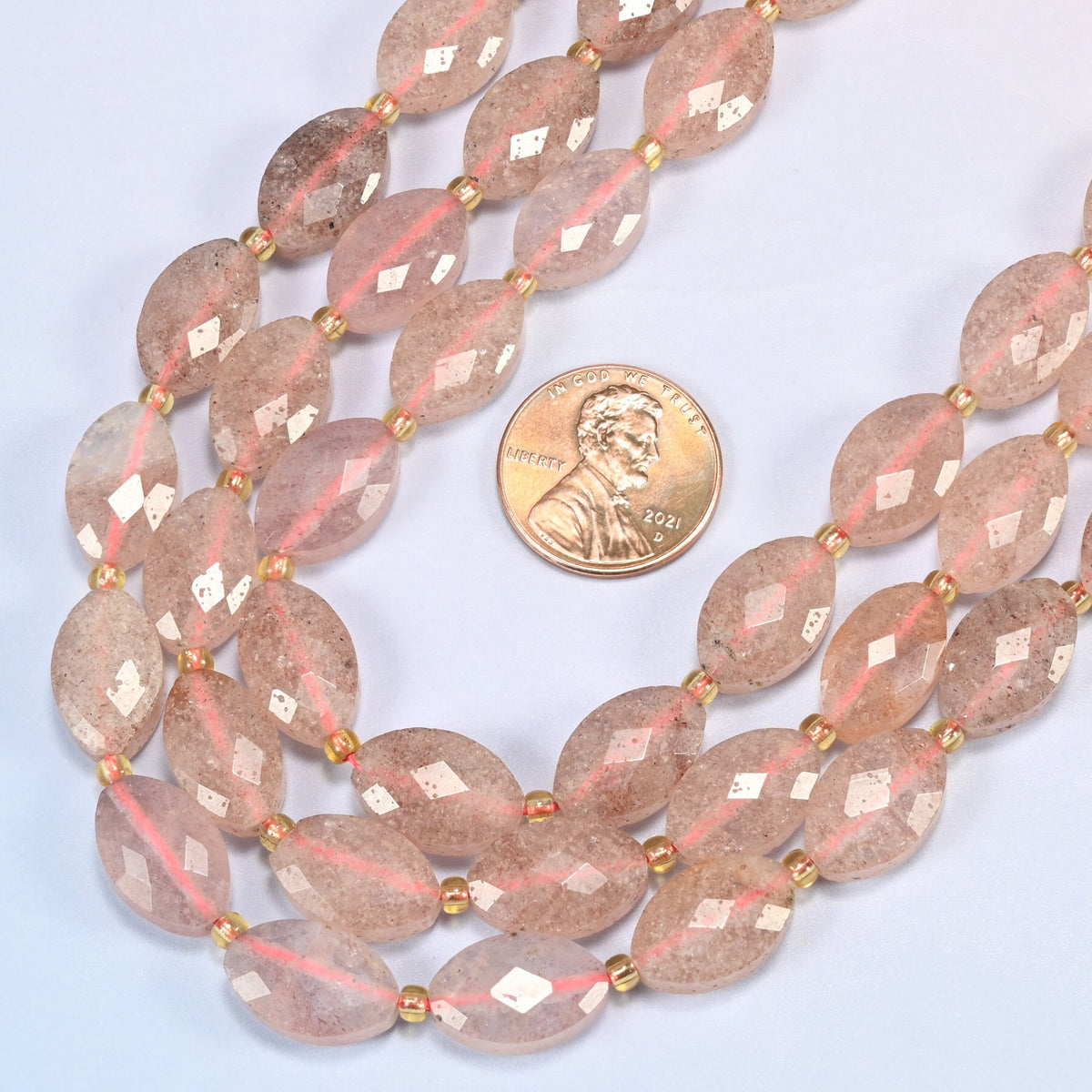 Close view of strawberry quartz faceted flat oval strands curving beside a US penny for scale