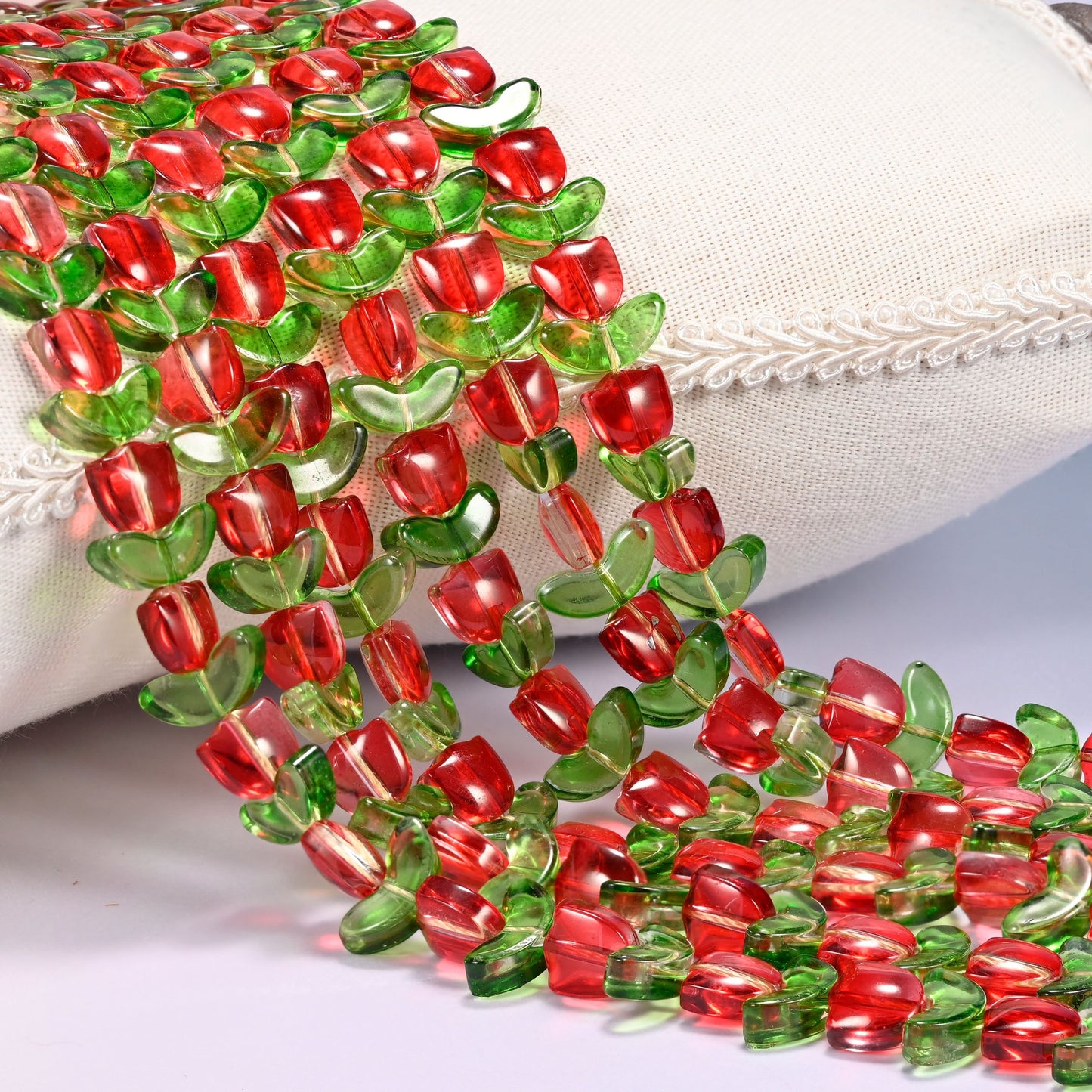 Close-up of red tulip flower and green leaf glass beads cascading diagonally over a cream woven pillow with decorative trim.