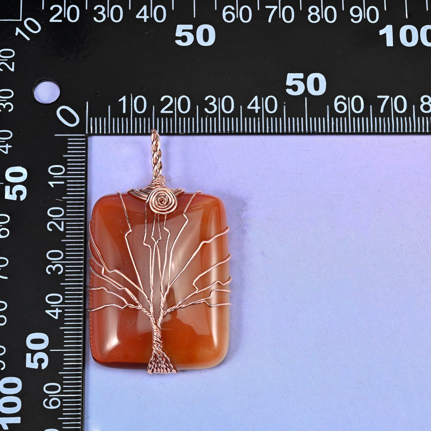 Rectangle Gemstone Tree of Life Pendant held against a black millimetre ruler for scale, the red-orange piece measuring about 55x30mm.